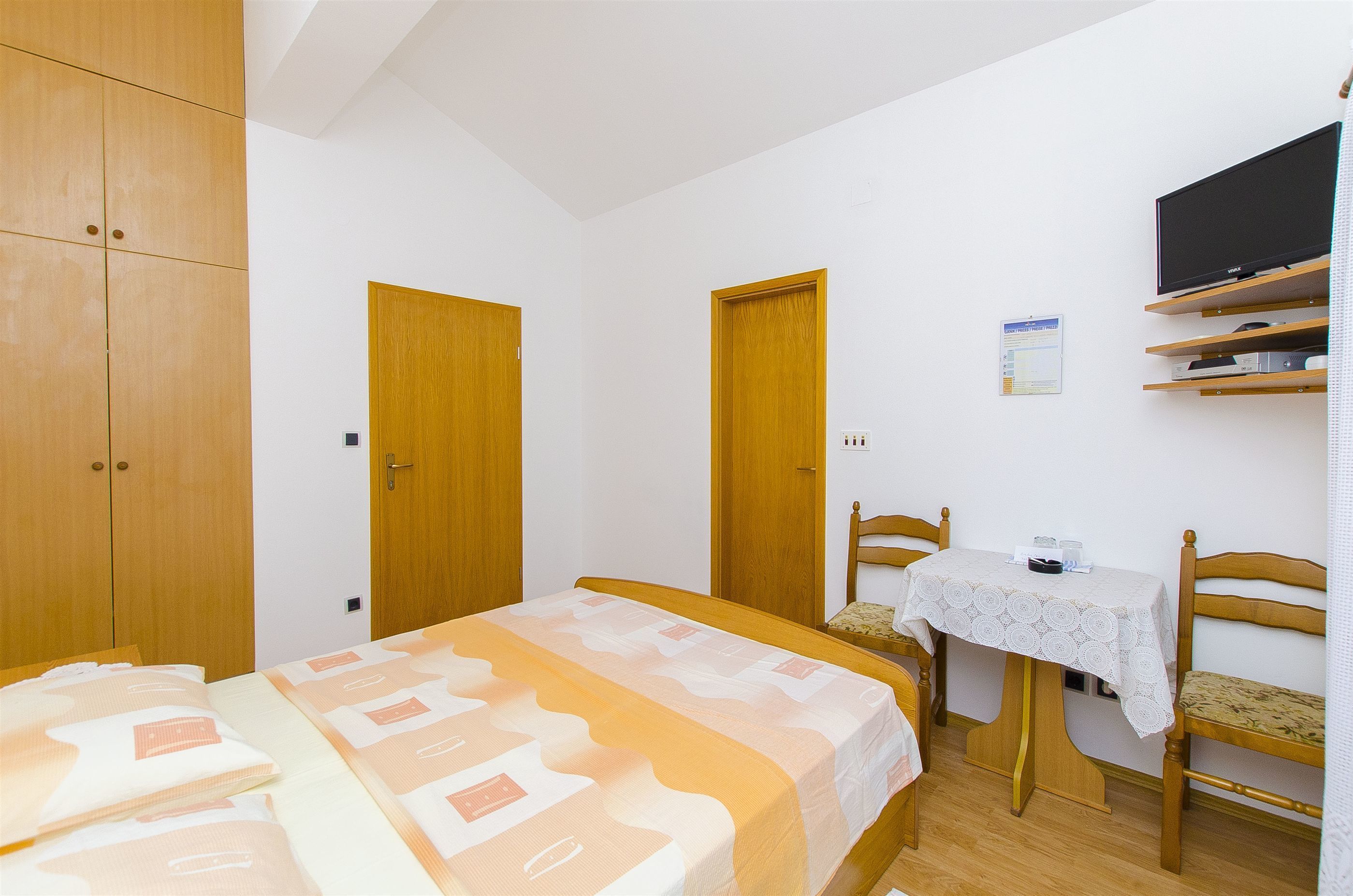 A bedroom with a bed, a table with a chair, wooden wardrobes, and a mounted TV.