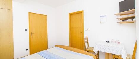 A bedroom with two wooden doors, a bed with a patterned bedspread, a small table with a white lace tablecloth, and a chair.