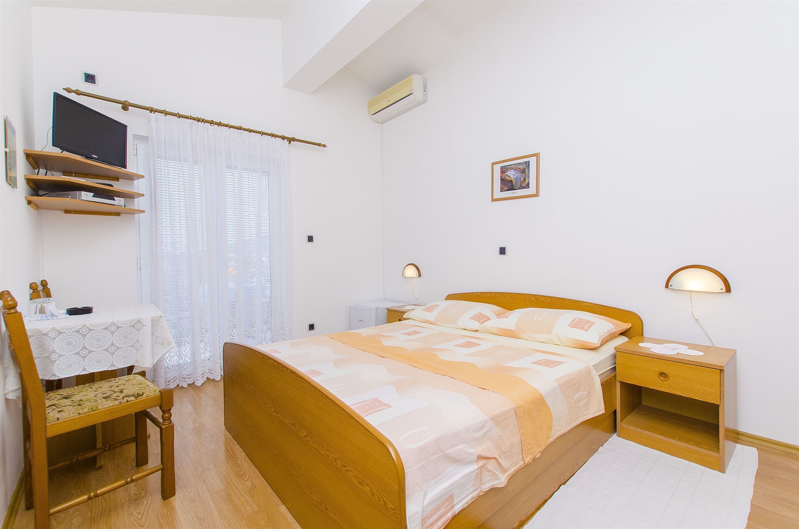 A bedroom with a wooden bed, a bedside table with a lamp, a small bedside table with a lamp, a desk with a chair, a window with curtains, and a wall-mounted air conditioner.