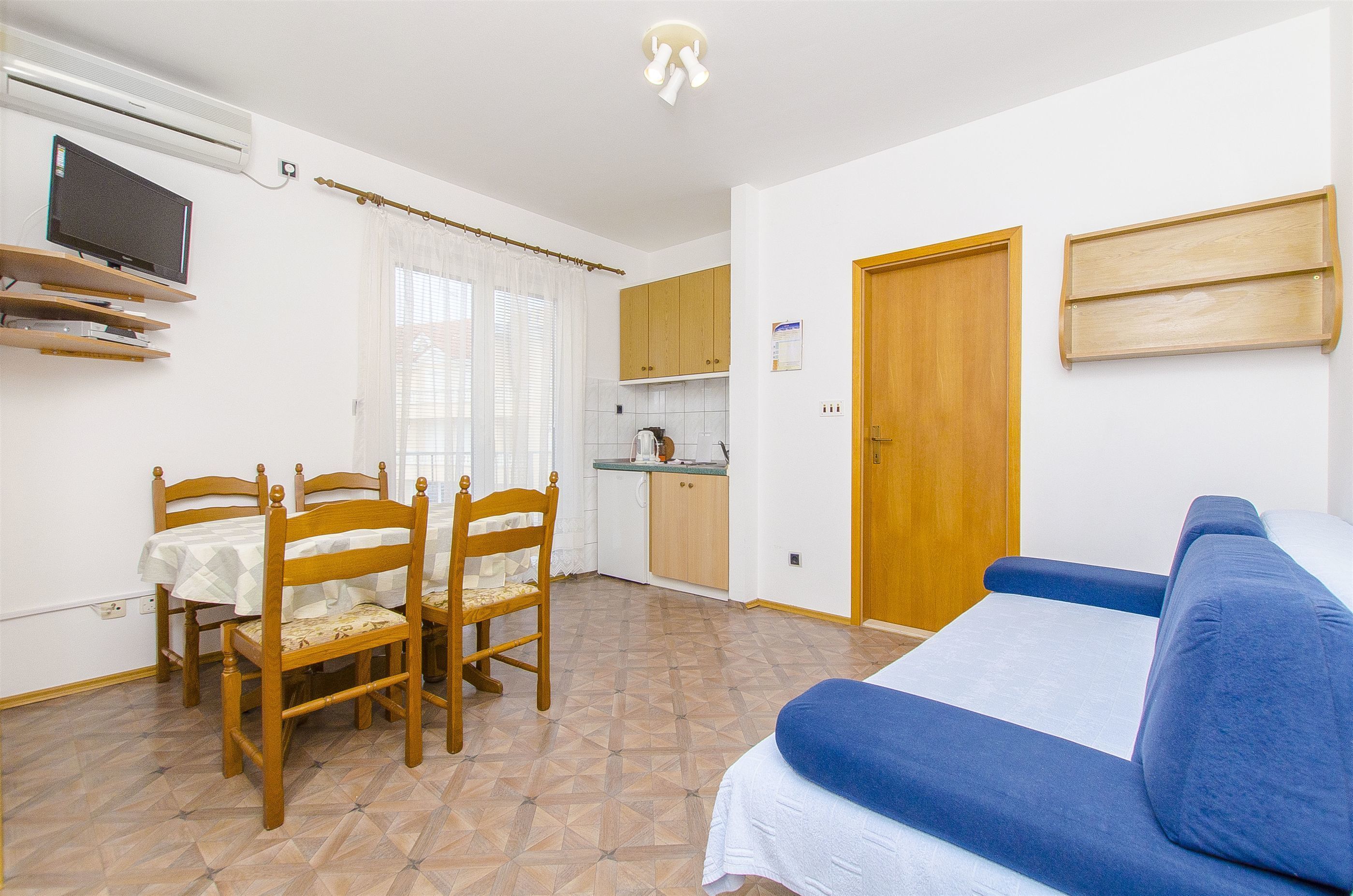 A compact room with a dining table, chairs, a sofa, a kitchenette, and a wooden door.