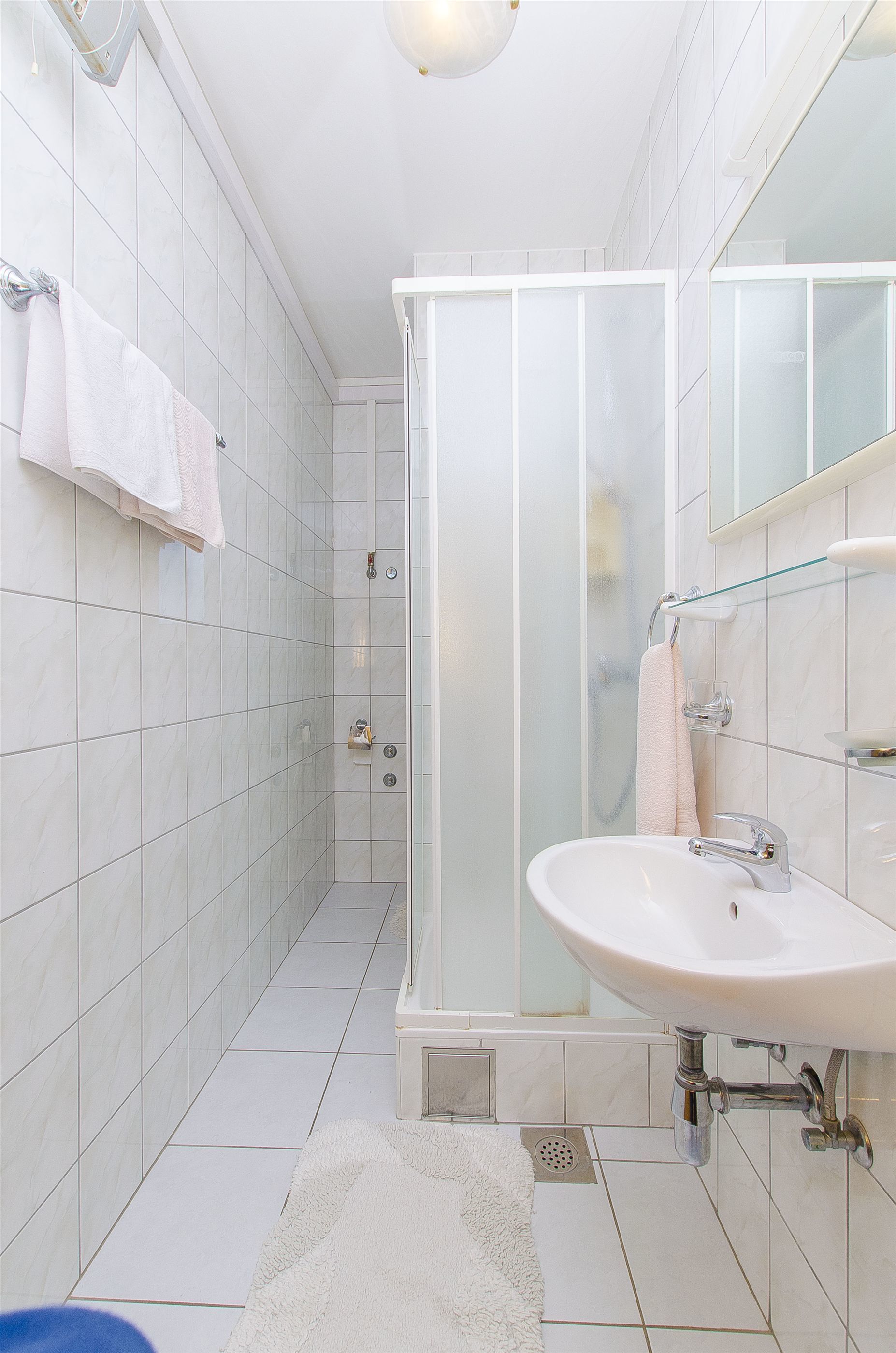 Un baño moderno con baldosas blancas, cabina de ducha de vidrio, lavamanos con espejo y toallero.
