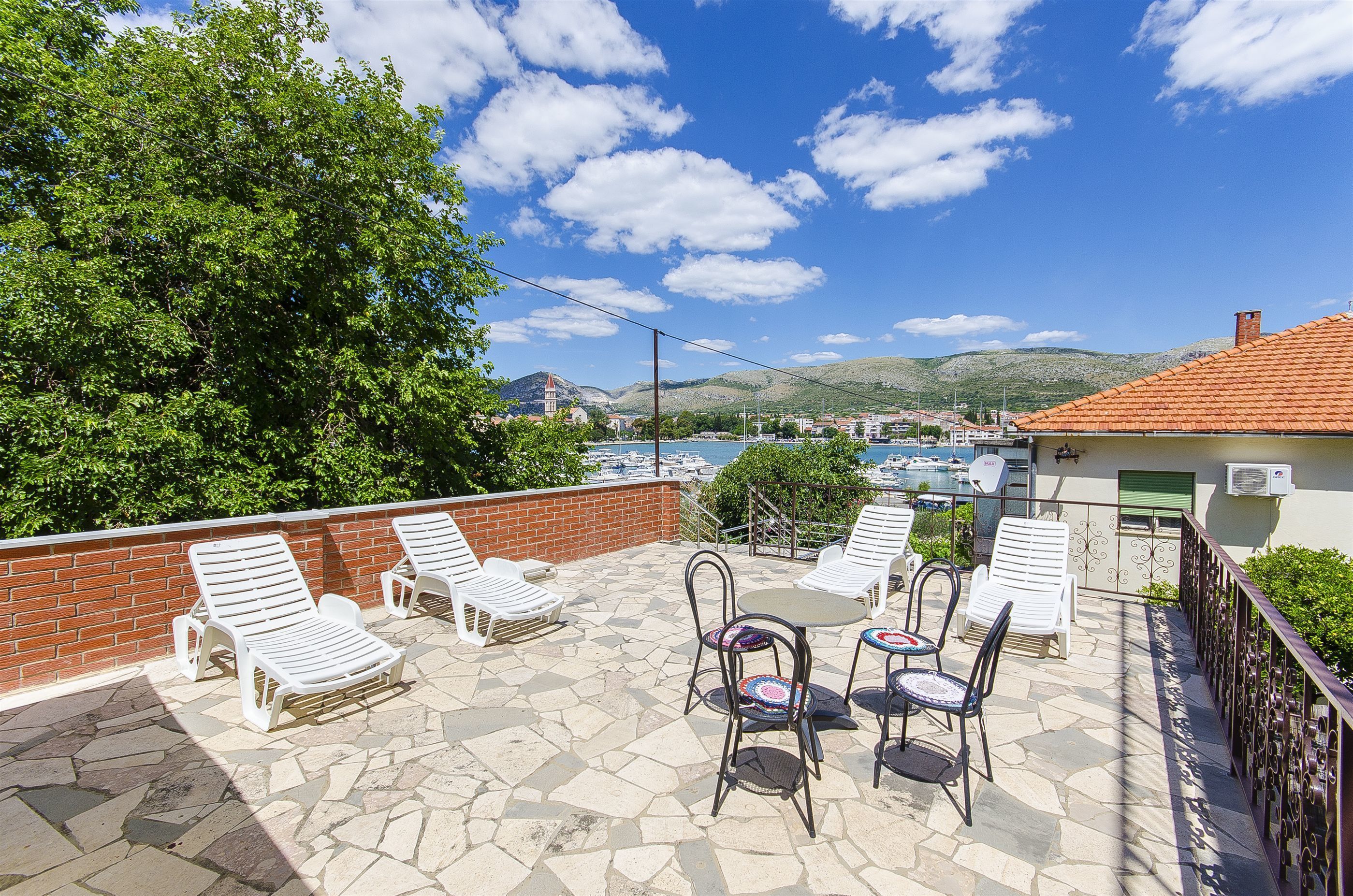 A terrace with stone flooring, white lounge chairs, a table with chairs, and a view of a marina and hills in the background.