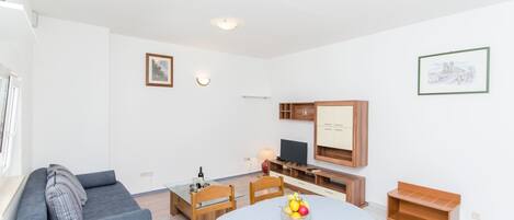 A modern living room with a grey sofa, a dining table with a fruit bowl, a TV, and wooden cabinets.