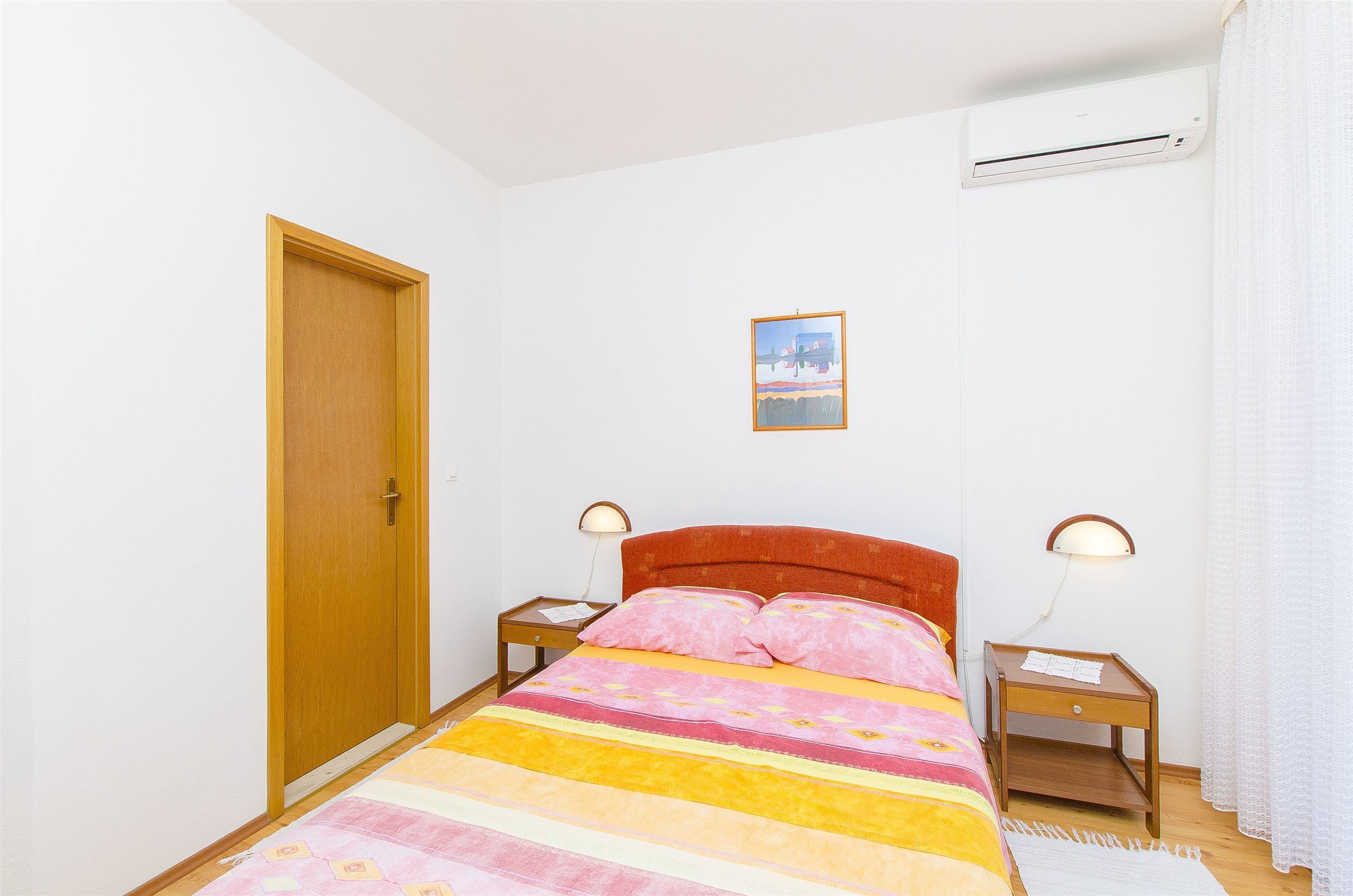 Una camera da letto con un letto, due comodini, un mobiletto e un condizionatore d'aria a parete, una porta e un quadro appeso al muro.
