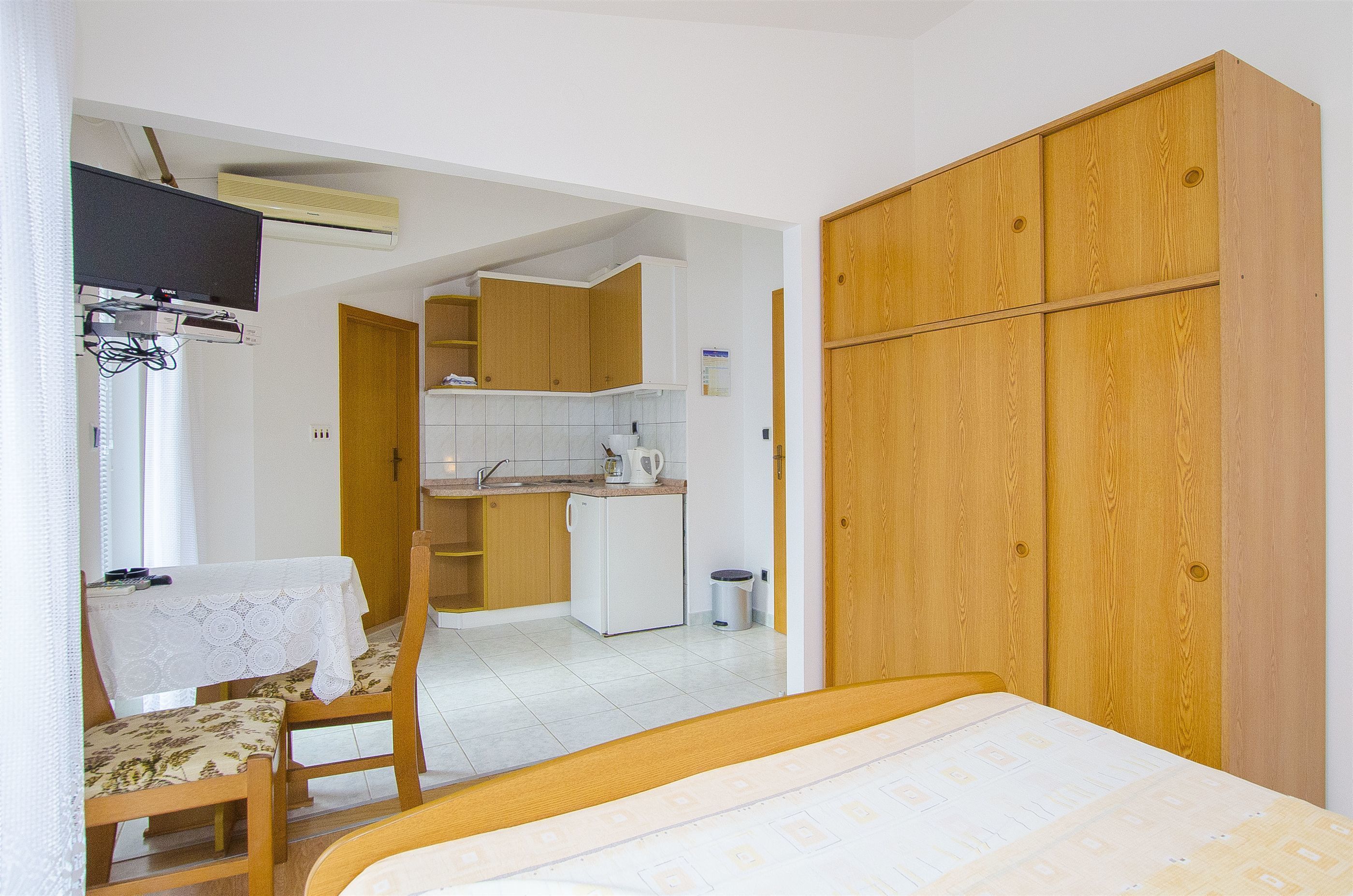 A compact bedroom with a bed, a wooden wardrobe, a small kitchen area, and a dining table with chairs.