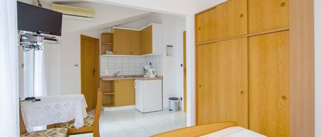 A compact bedroom with a bed, a wooden wardrobe, a small kitchen area, and a dining table with chairs.