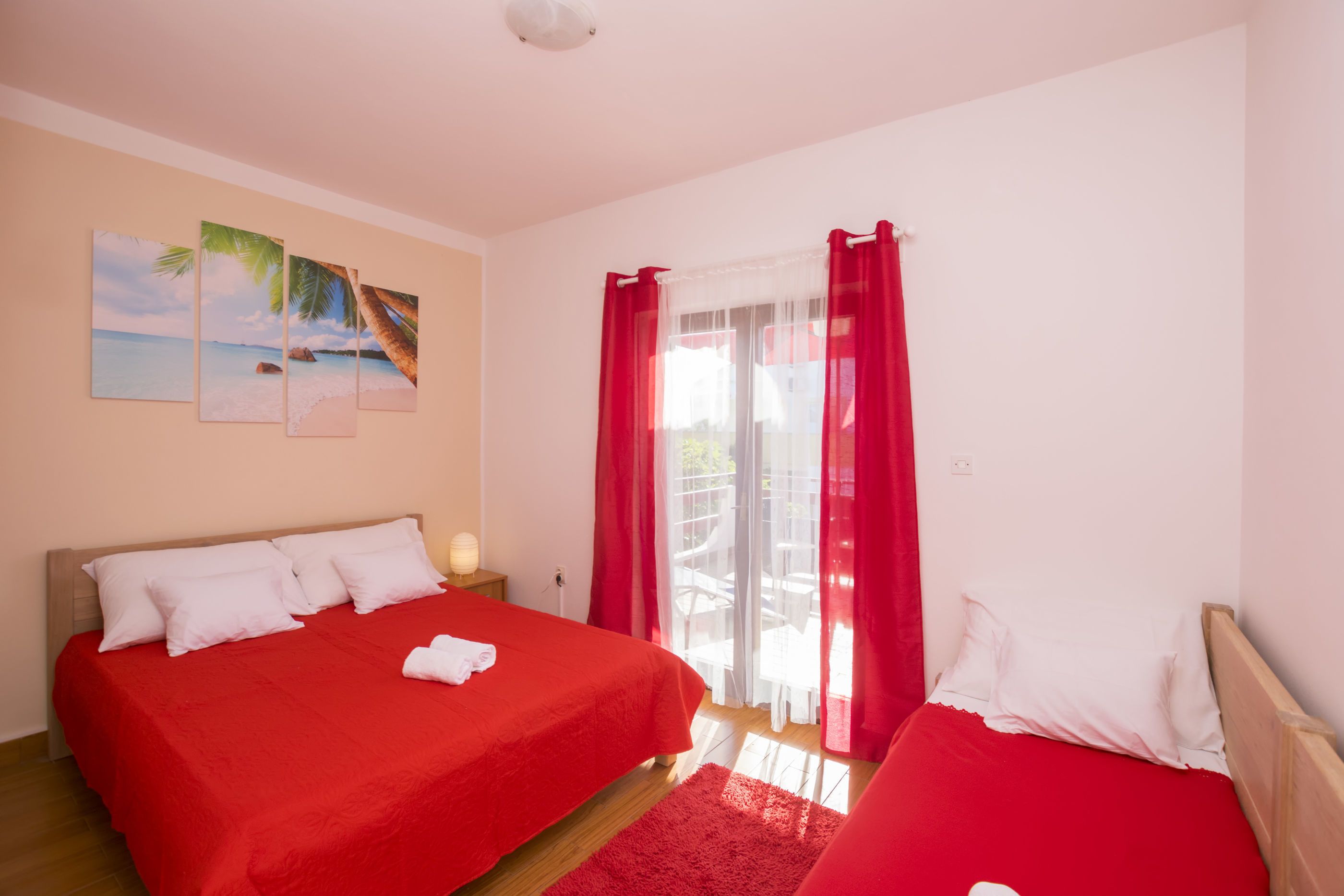 Een hotelkamer met twee bedden, een rode sprei, een rood gordijn, een balkon en een kunstwerk op de muur met een strandthema.