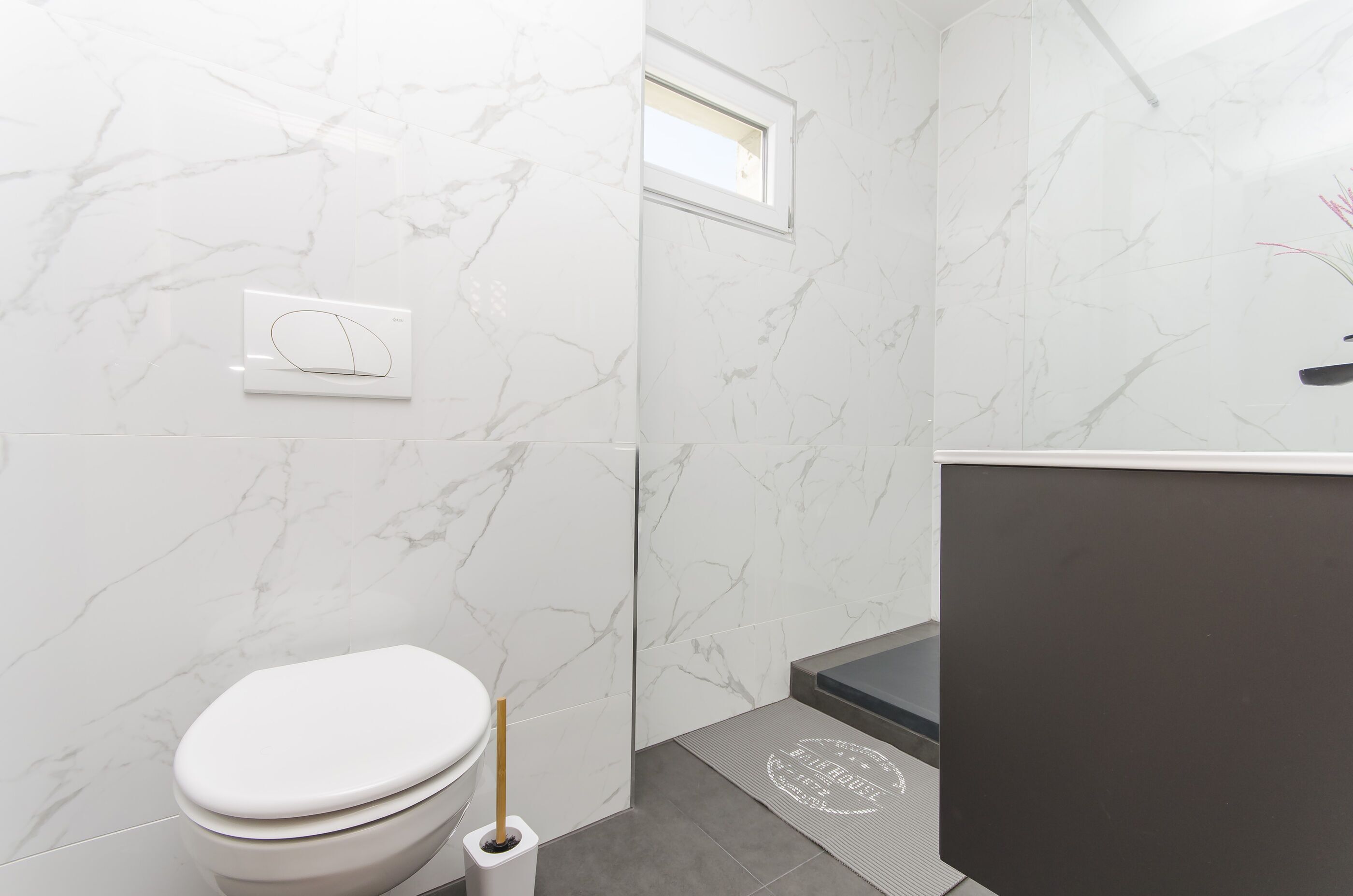 A modern bathroom with white marble walls, a toilet, a shower area, and a small window.