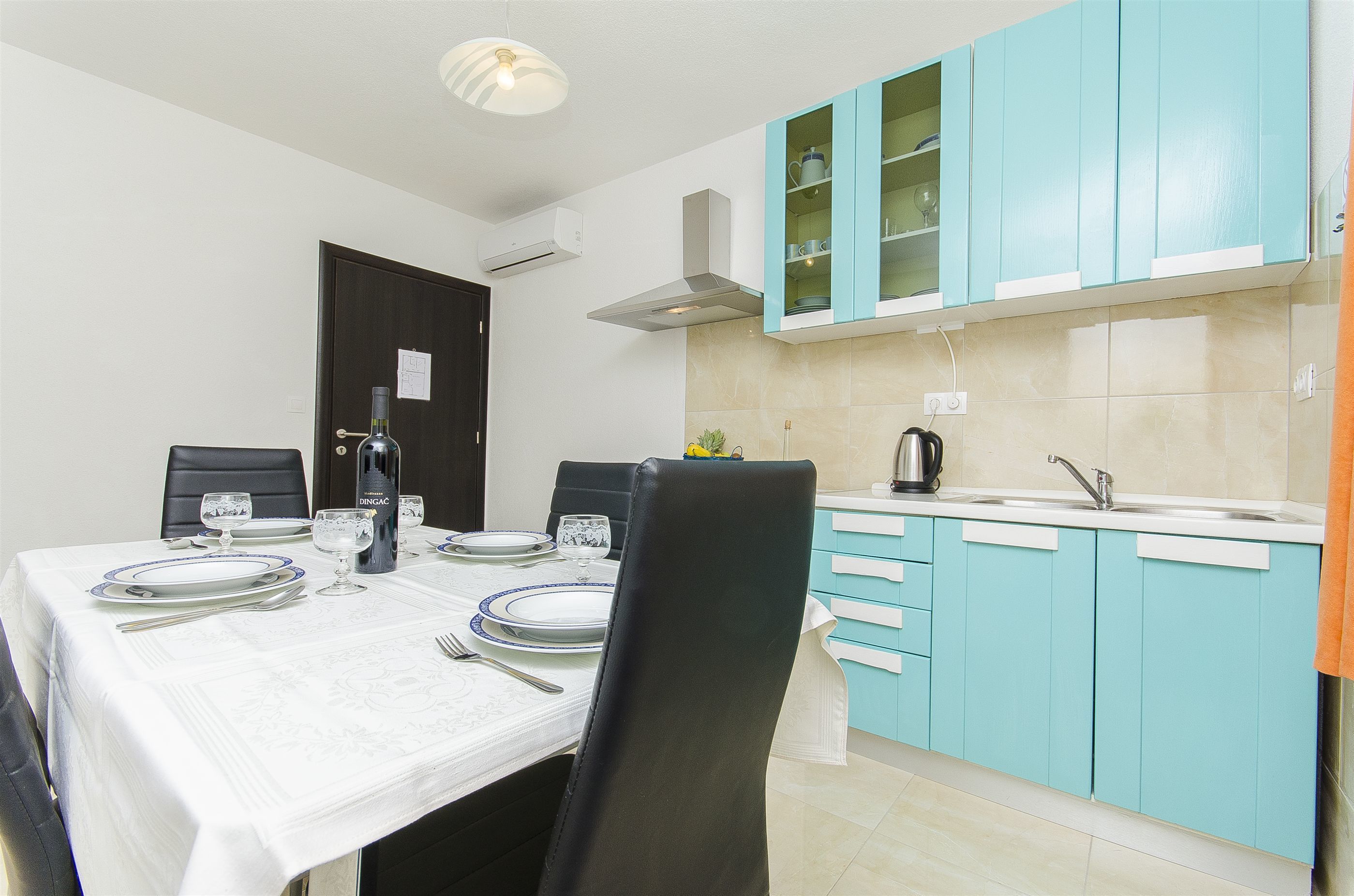 A dining area with a table set for four, a kitchen with teal cabinets, and a black chair in the foreground.