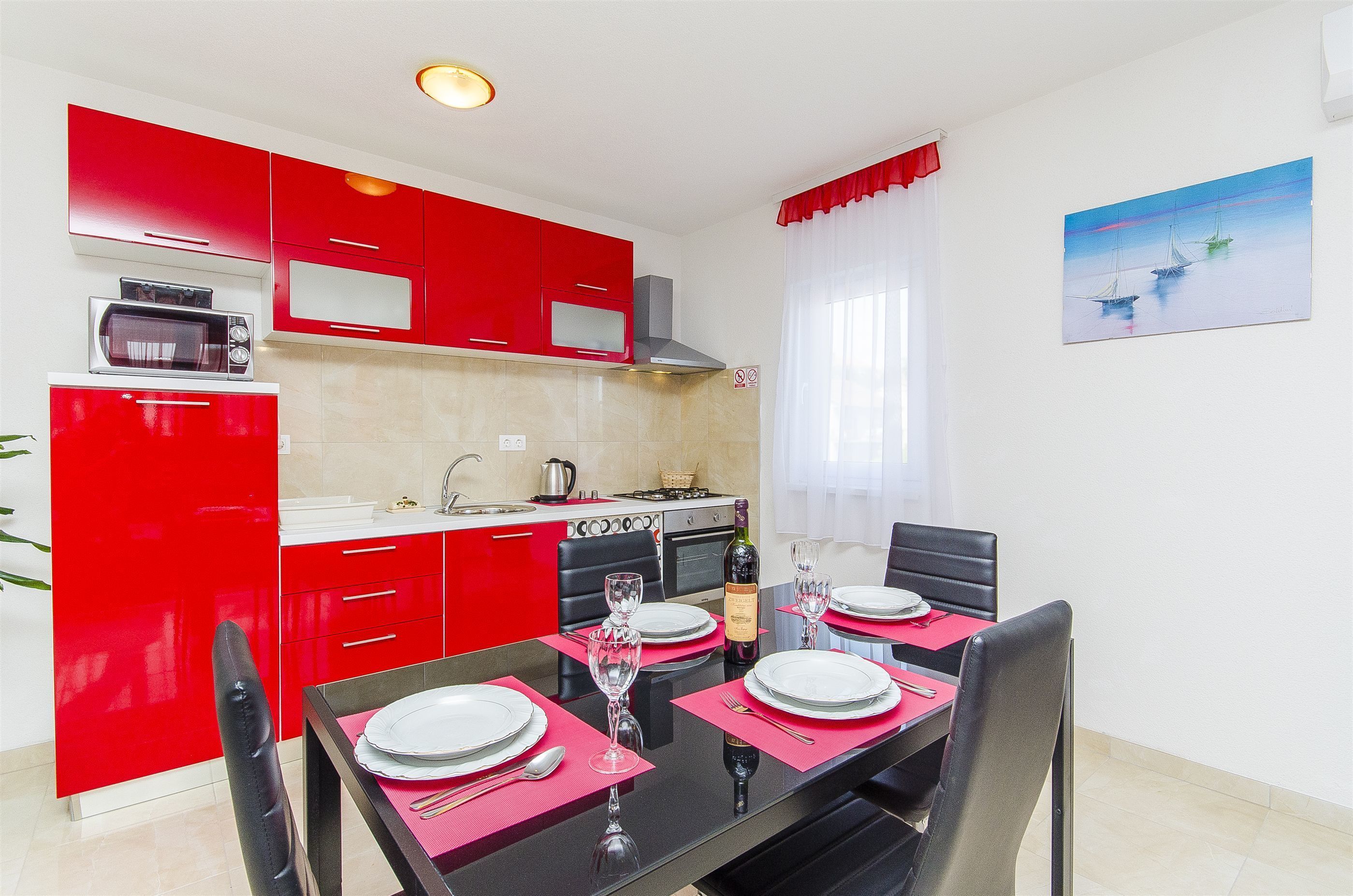 Una cocina moderna con armarios rojos, una mesa de comedor preparada para cuatro personas y un cuadro de veleros en la pared.