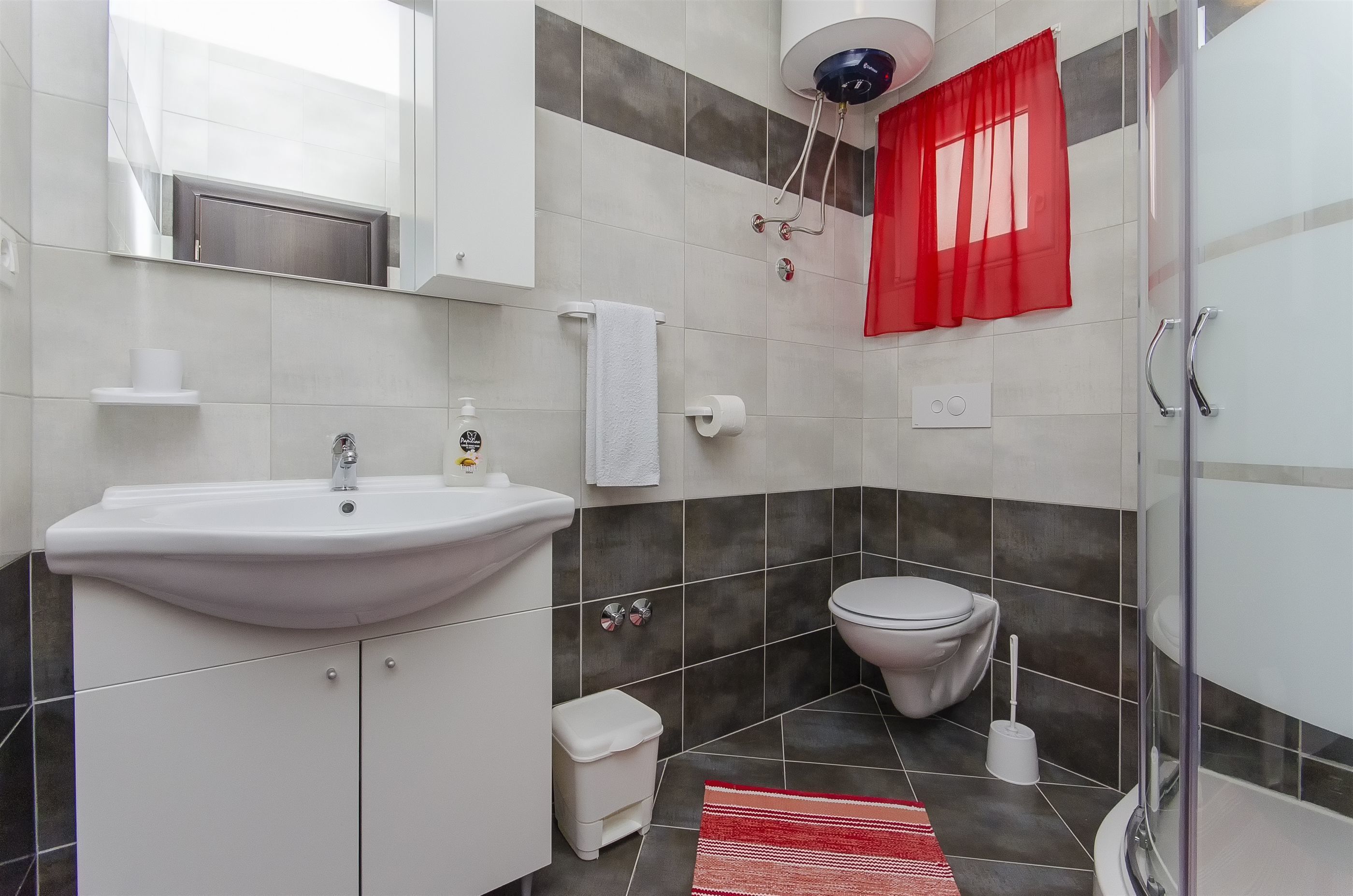 A bathroom with a white sink, toilet, and shower. There is a red curtain and a white towel hanging on the wall.