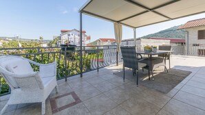 Un balcón techado al aire libre con una mesa y sillas, una silla de mimbre y vistas a edificios y vegetación.