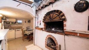 A kitchen with a built-in wood-fired oven, a dining area with a table and chairs, and a white tiled floor. - Apartment Ivan Vrsi (Vrsi)