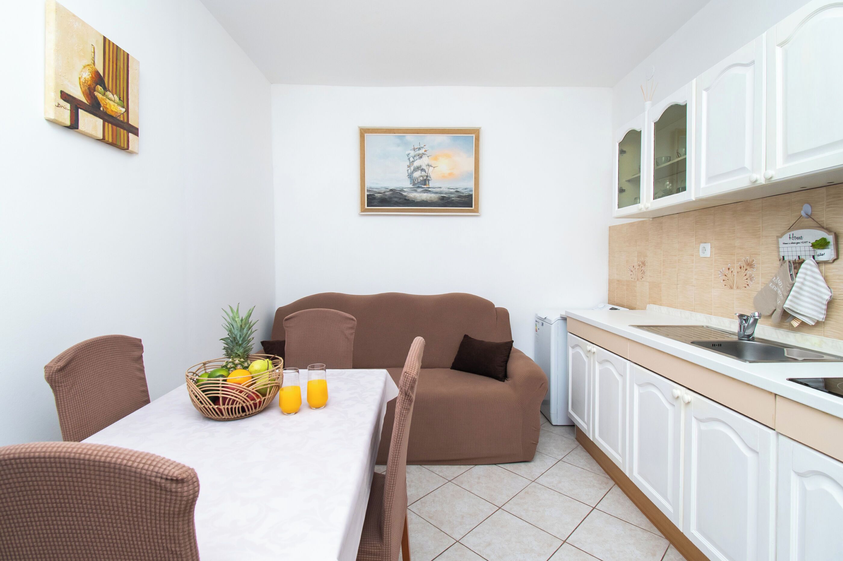 Eine kompakte Küche mit weißen Schränken, ein Essbereich mit Tisch und Stühlen sowie ein braunes Sofa.
