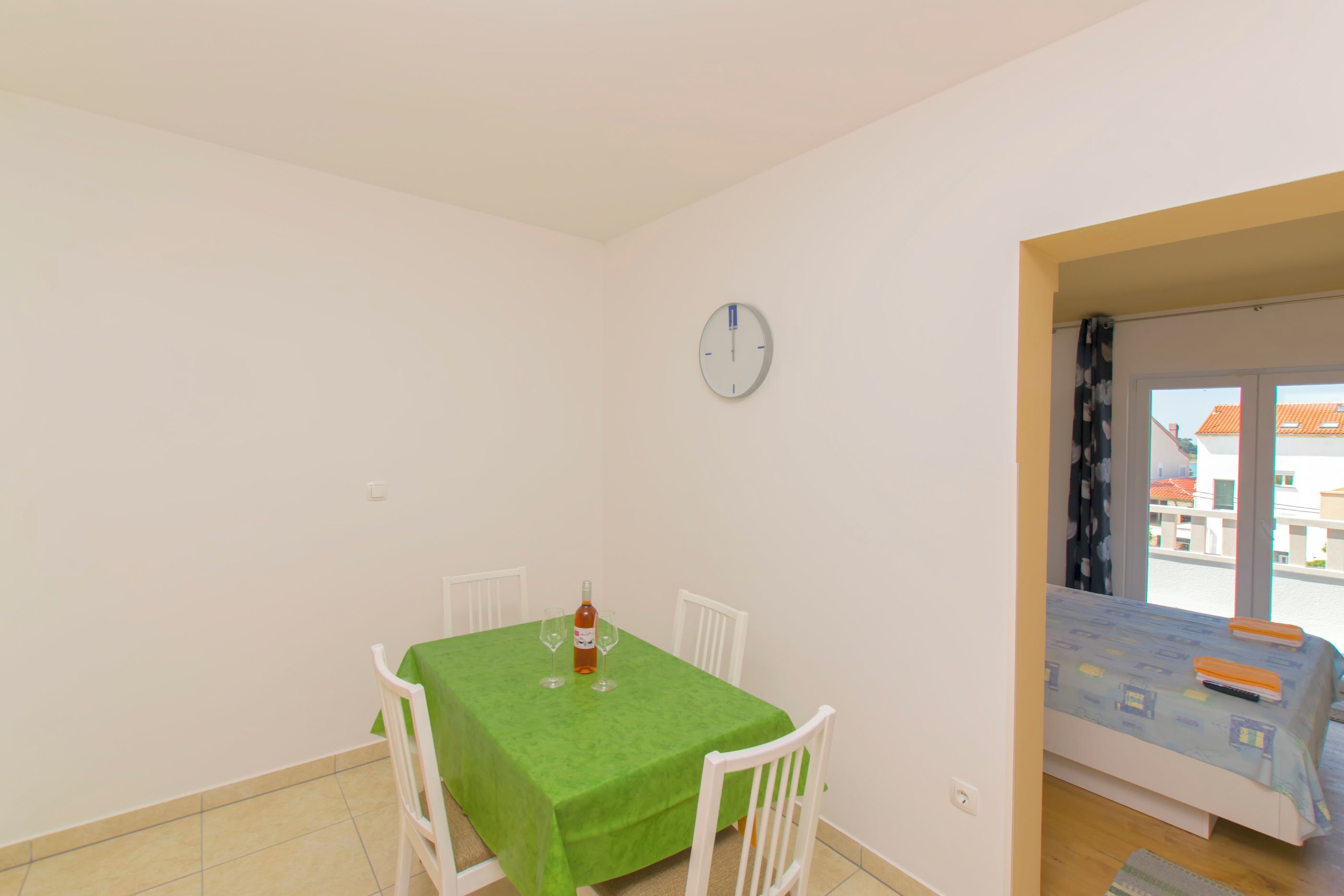 A room with a dining table, chairs, a bed, a window, and a wall clock.
