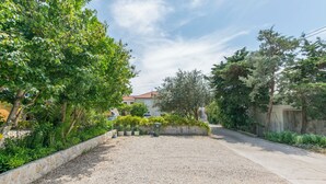 A residential area with a gravel driveway, a stone wall, and various trees. - Apartments and Room Central Position (Novalja)