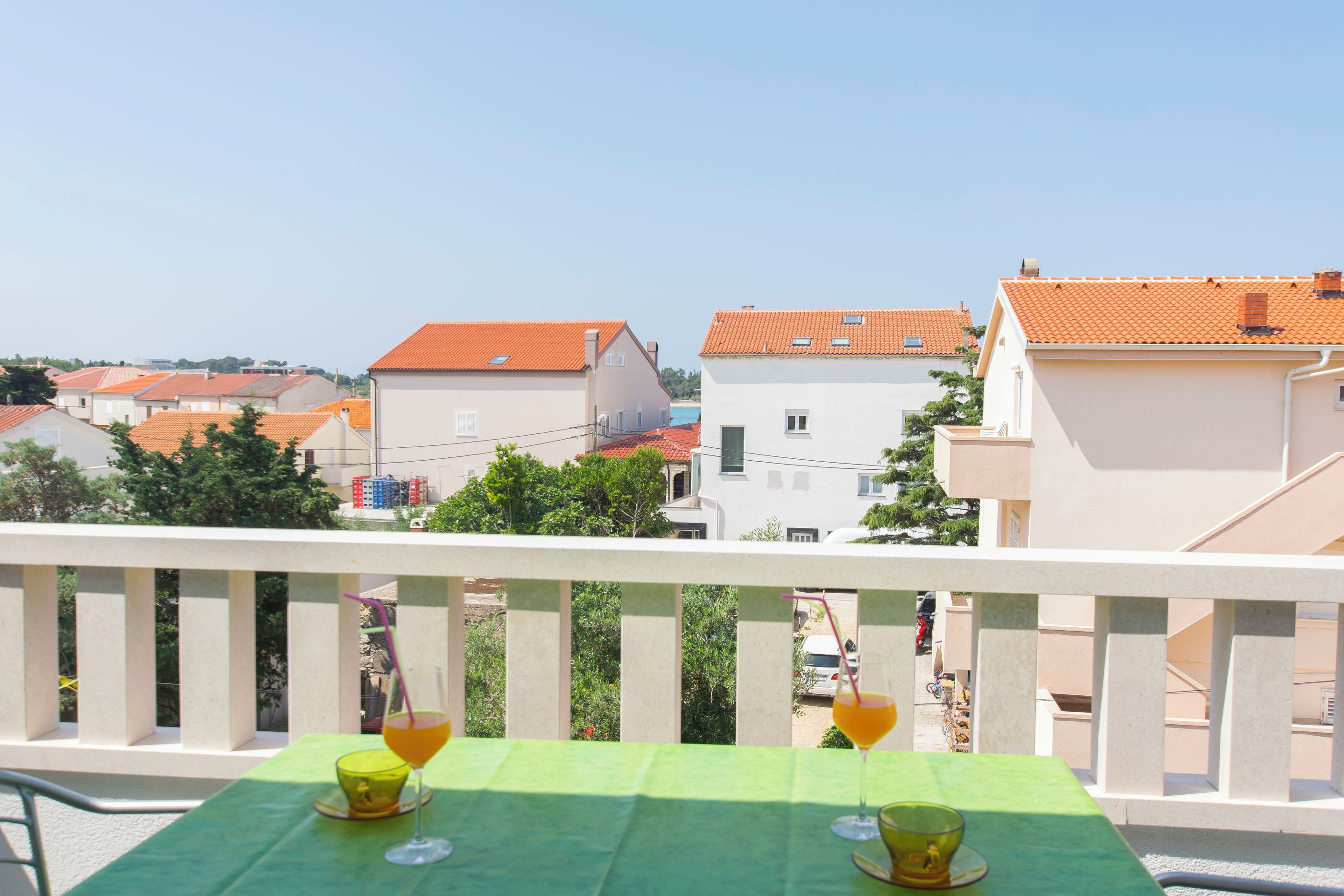 A balcony with a green tablecloth, two glasses of orange drink, and a view of buildings with red roofs.