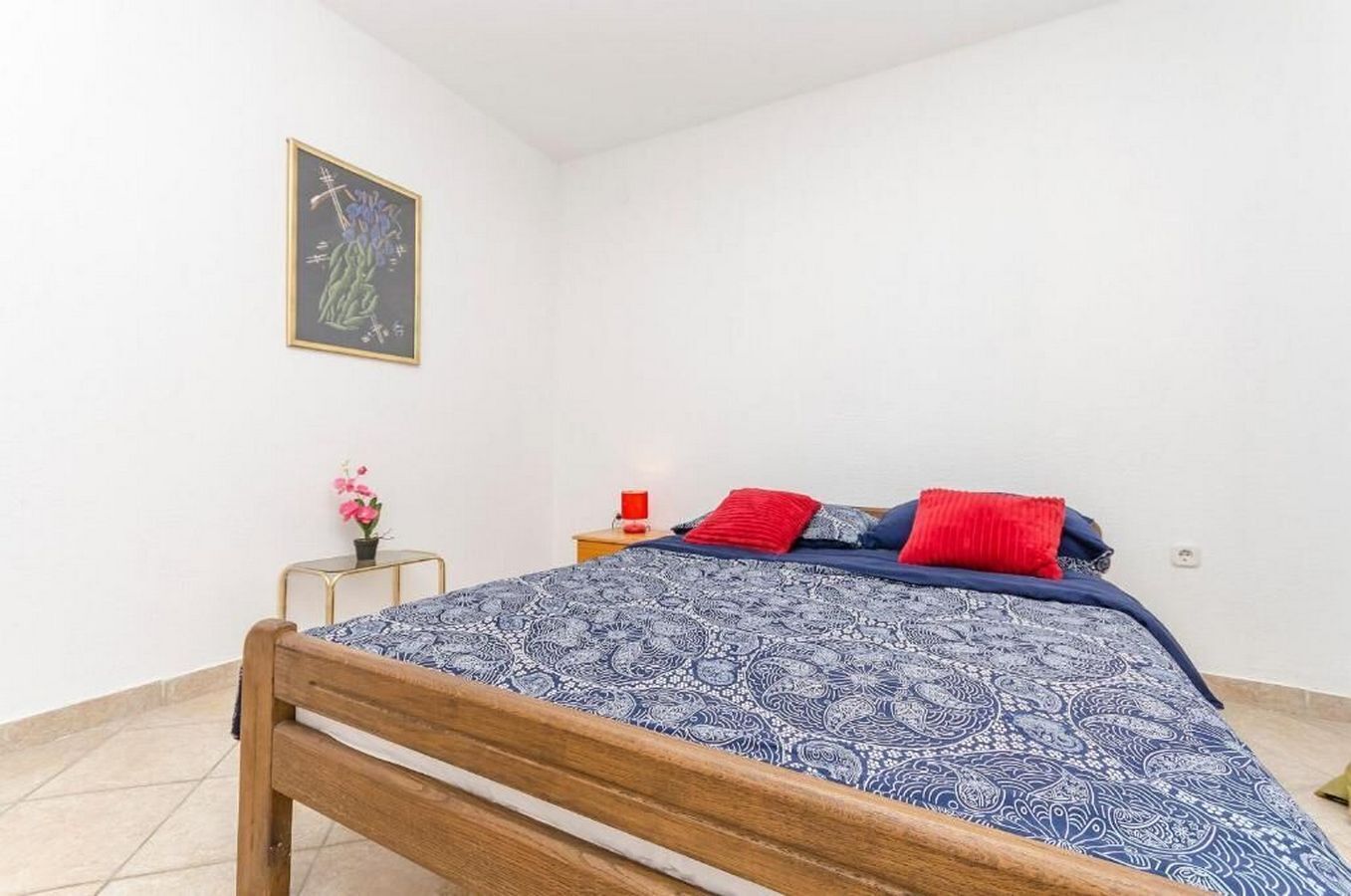 Une chambre avec un lit en bois, une table de chevet sur laquelle se trouve une lampe rouge, un tableau encadré accroché au mur et une petite table ornée d’une fleur rose.