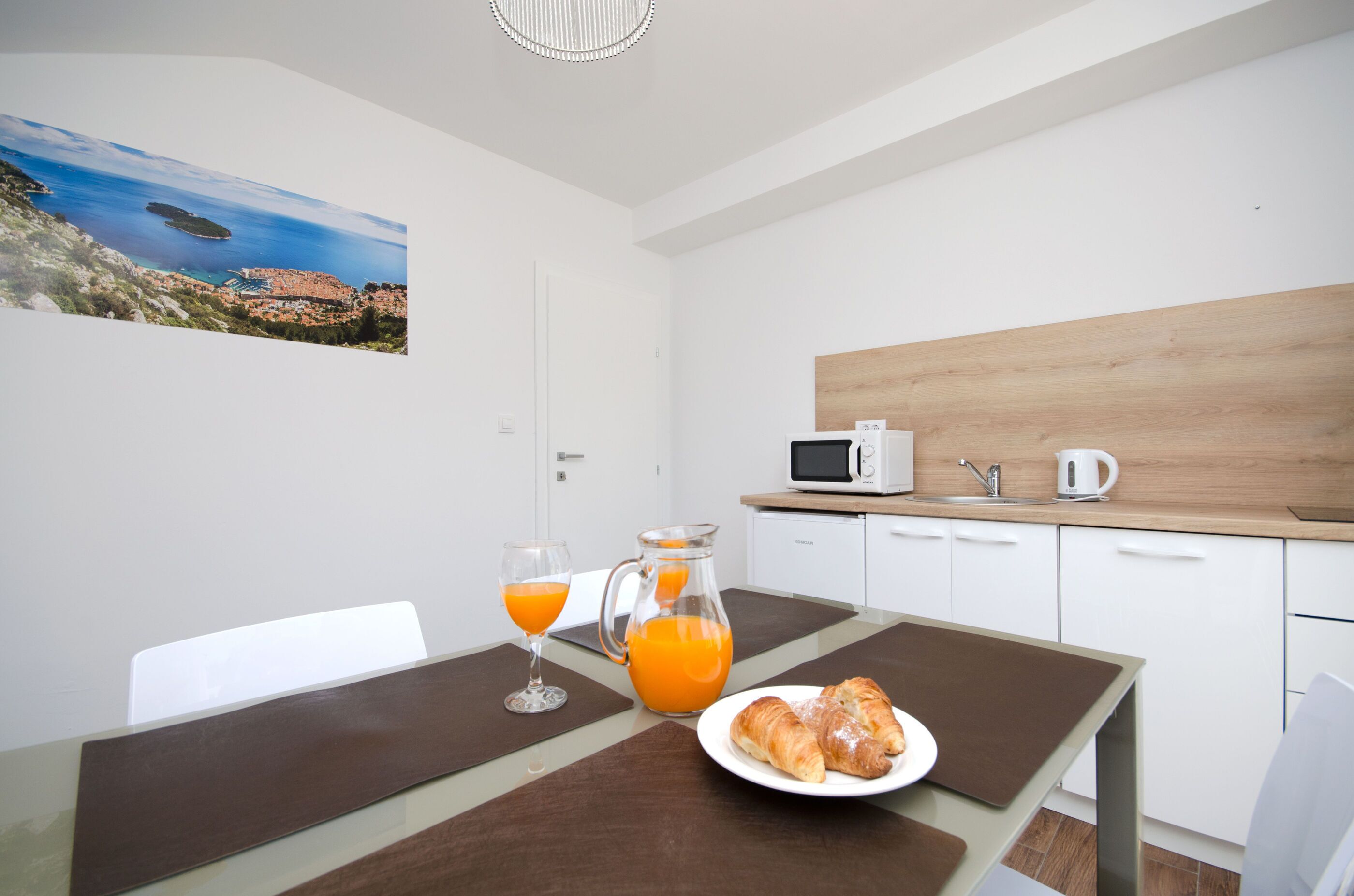 A modern kitchen with white cabinets, a wooden countertop, and a microwave. There is a table with a pitcher of orange juice and a plate with croissants.