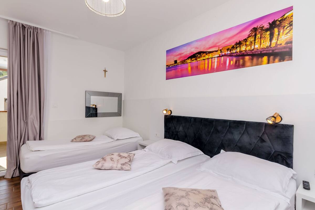 Hotelkamer met twee enkele bedden, een groot raam met gordijnen, een spiegel aan de muur en een schilderij van een stadsgezicht.