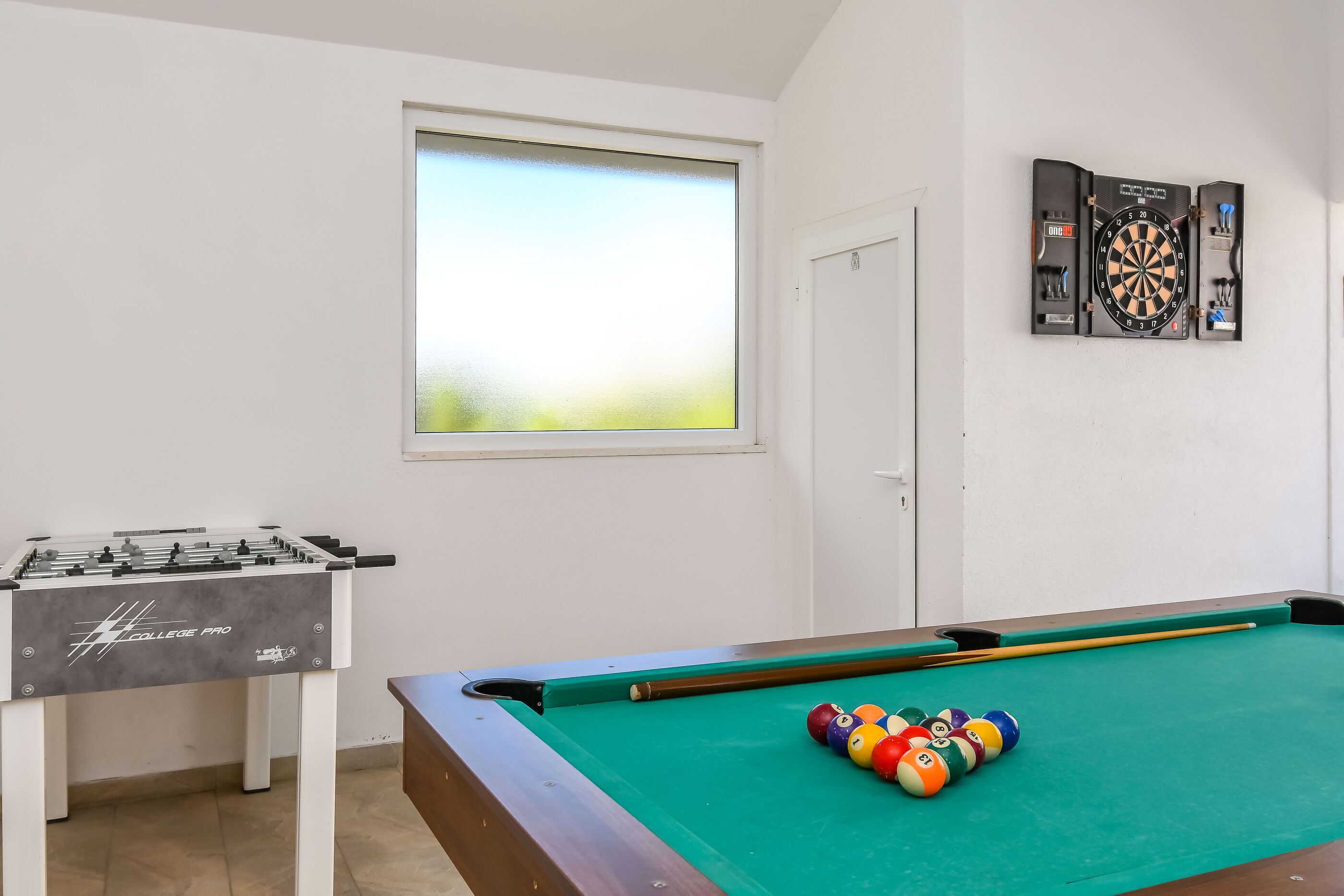 A room with a foosball table, a pool table, and a dartboard.