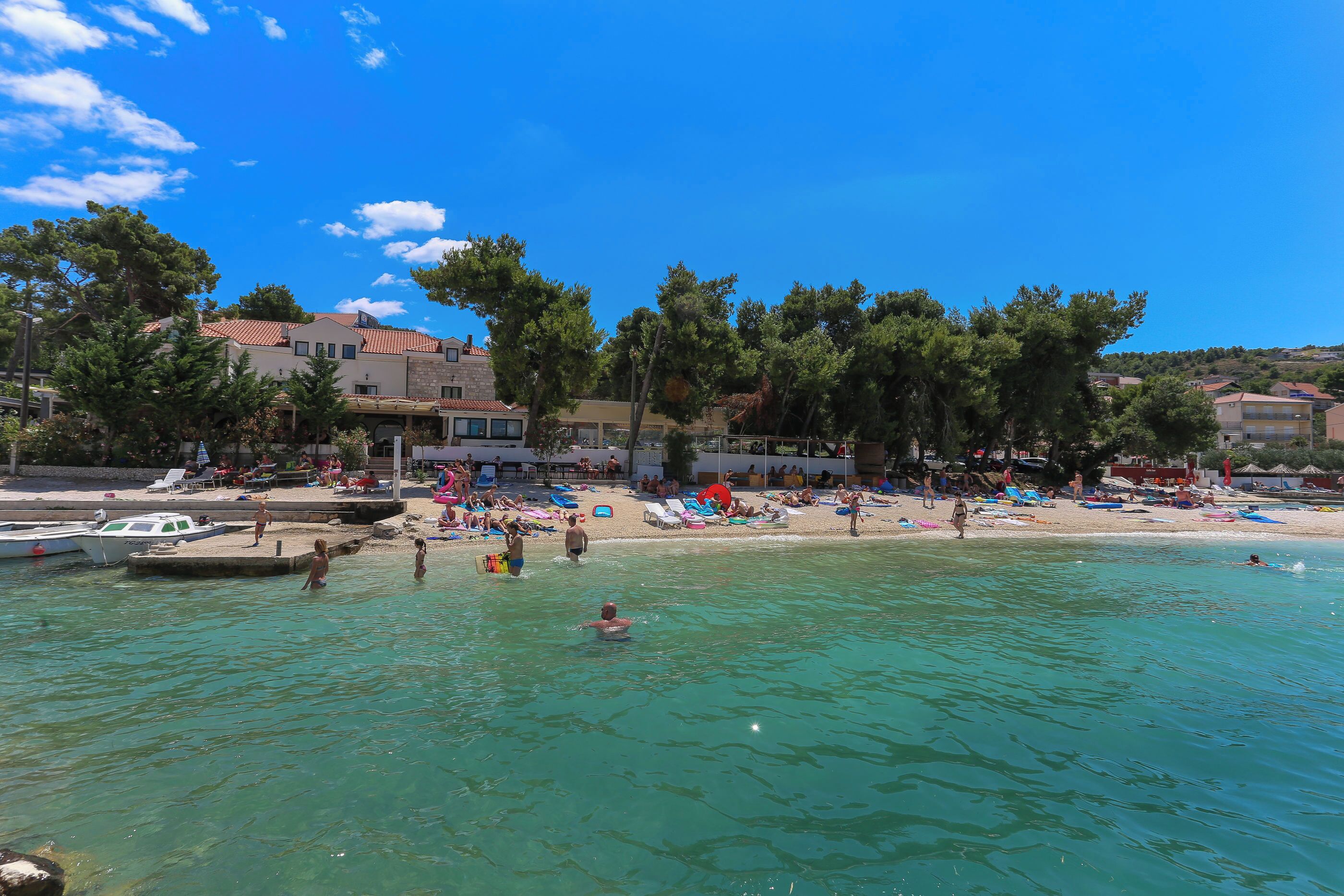 A beach with clear turquoise water, people swimming and sunbathing, and buildings in the background.