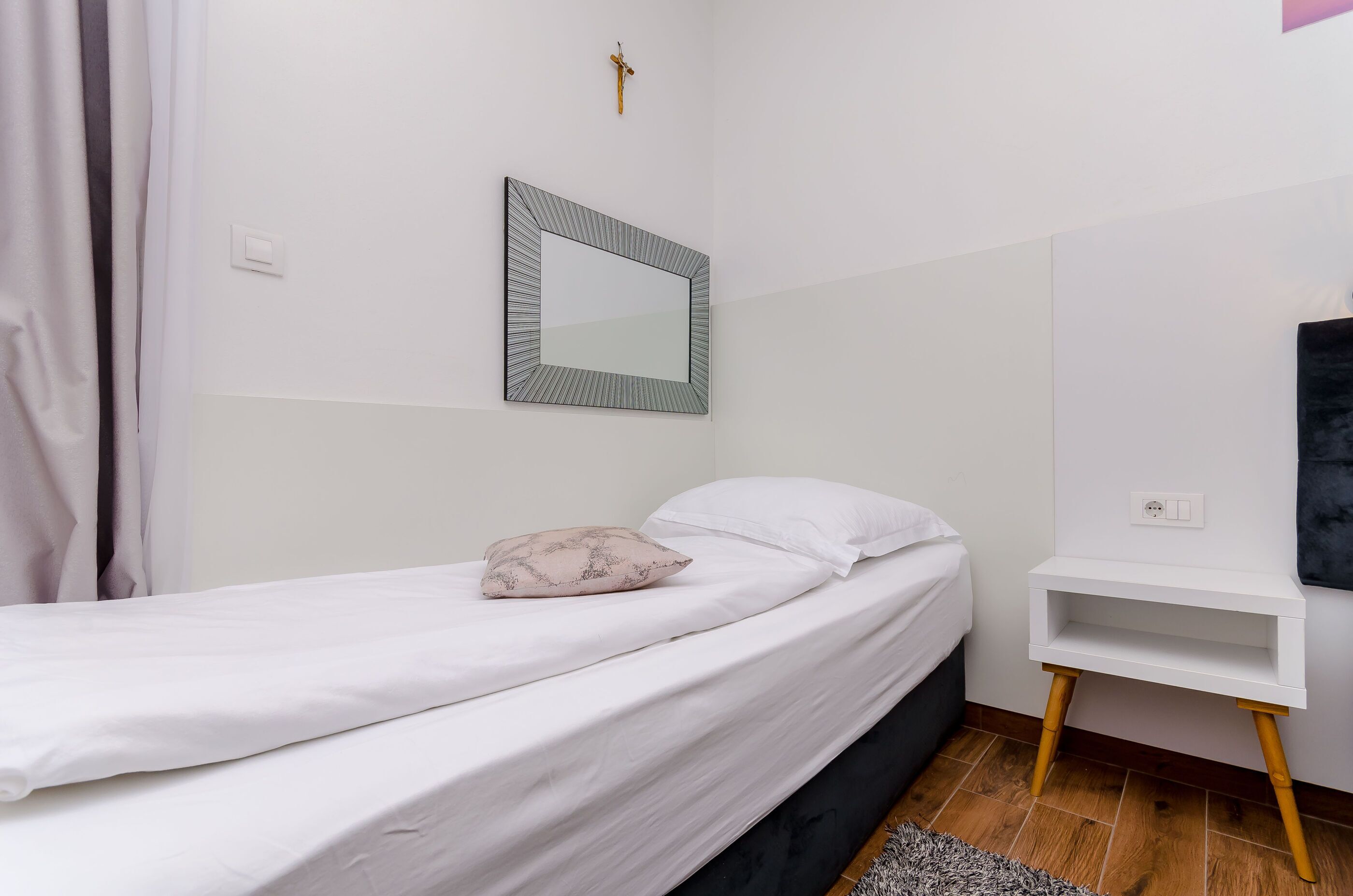A bedroom with a bed, a bedside table, a wall-mounted mirror, and a small shelf.