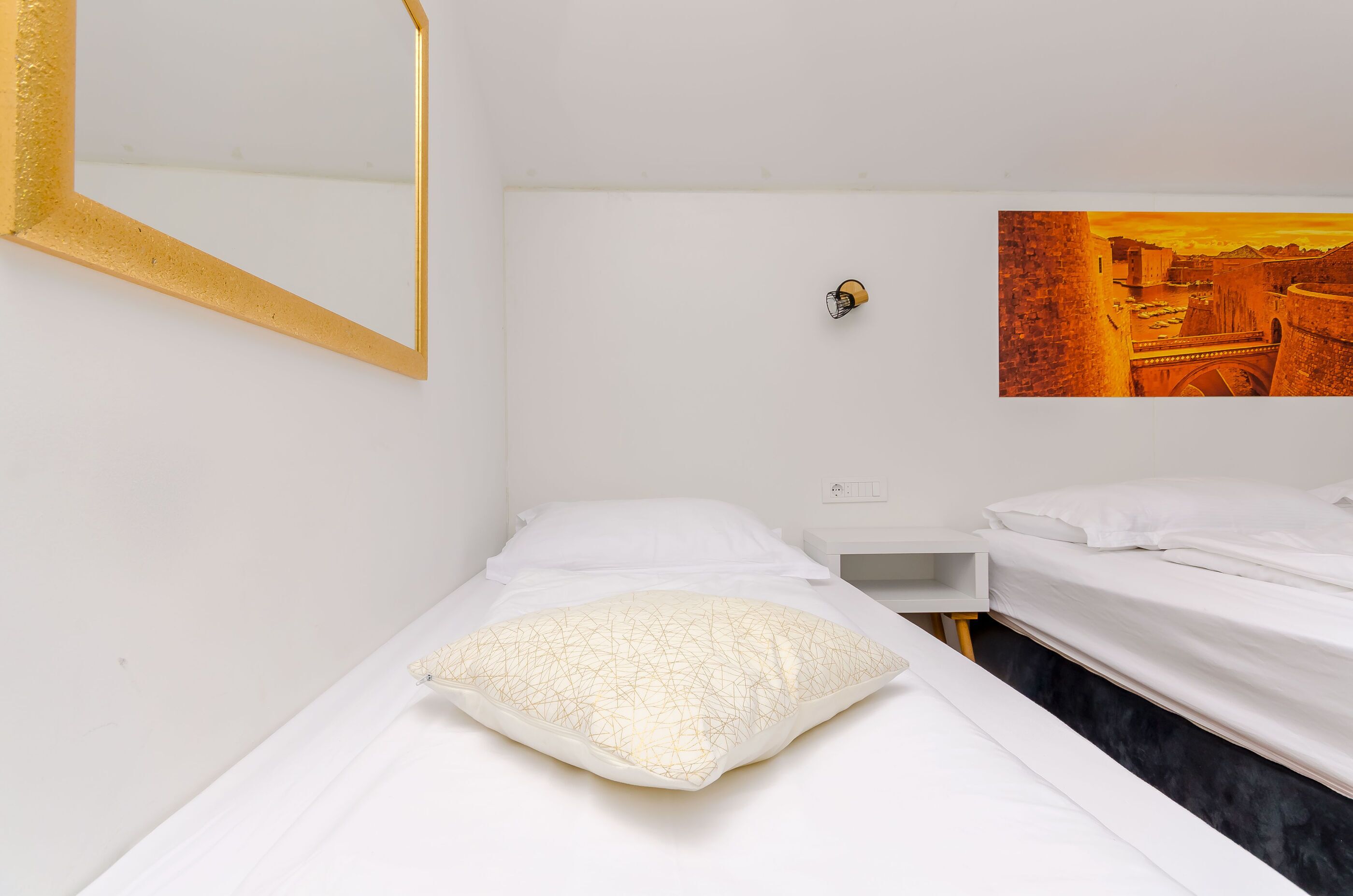 Un letto singolo con materasso bianco e un cuscino decorativo, un comodino, uno specchio fissato alla parete e un quadro che raffigura un paesaggio urbano.