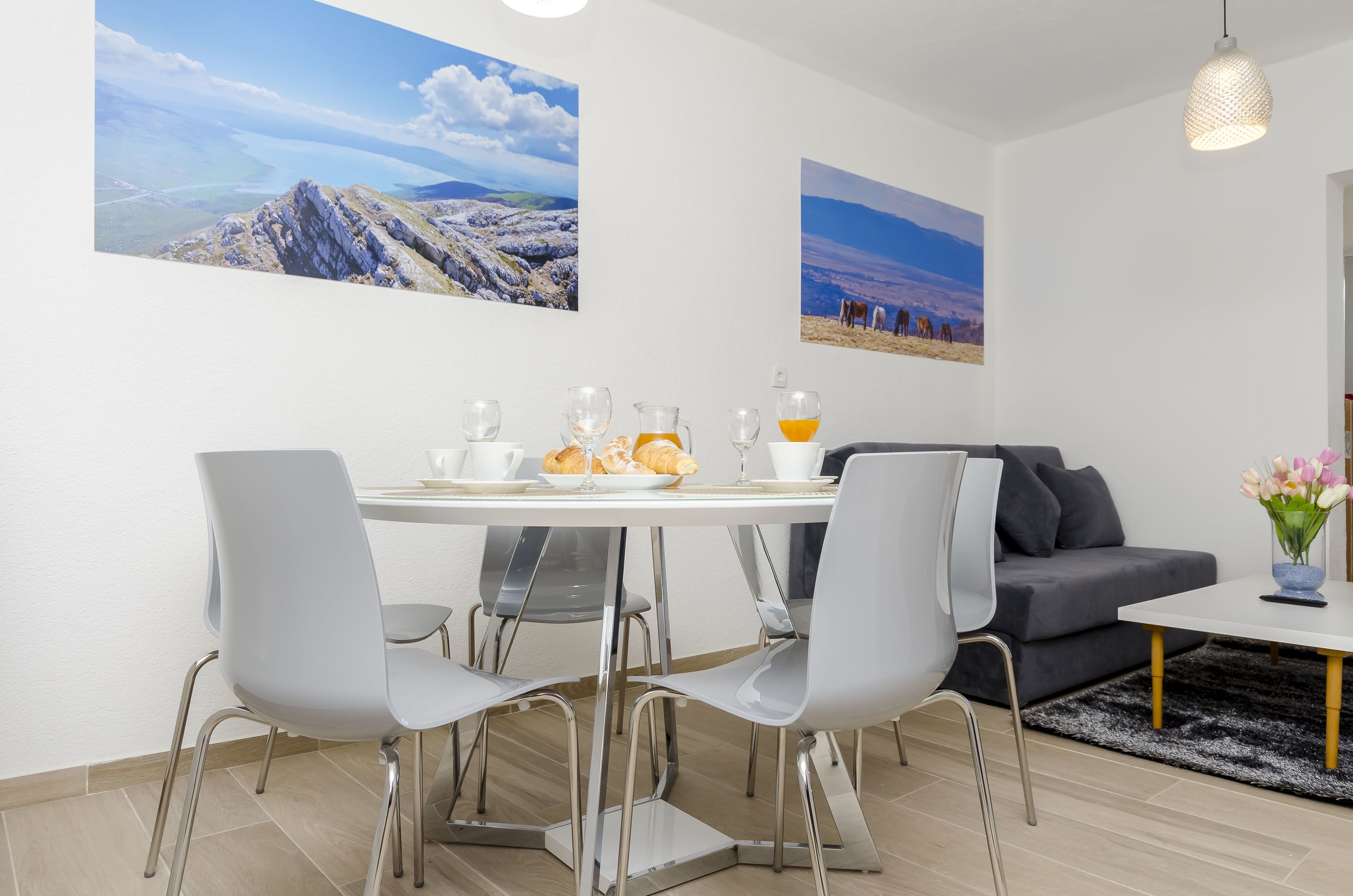 A modern dining area with a round table, grey chairs, and a grey sofa. There are two paintings on the wall, a black and white rug, and a hanging light fixture.