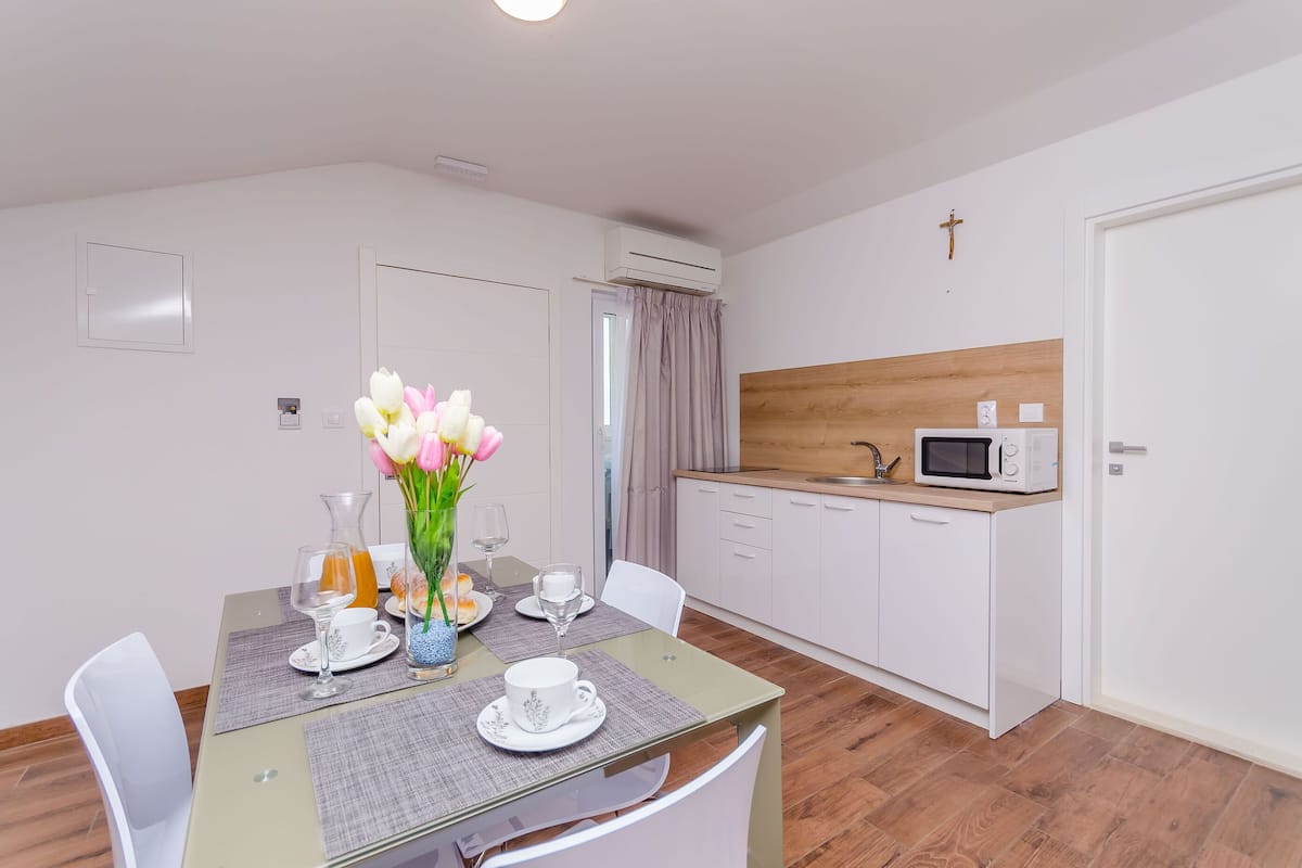 Een moderne eethoek met een glazen tafel, witte stoelen, een vaas met bloemen en een keuken met witte kasten en een magnetron.