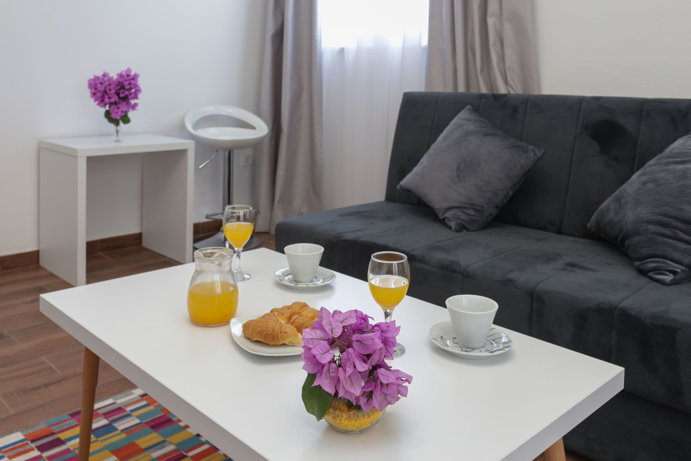 A modern living room with a white coffee table, a grey sofa, and a small white side table with a purple flower vase.