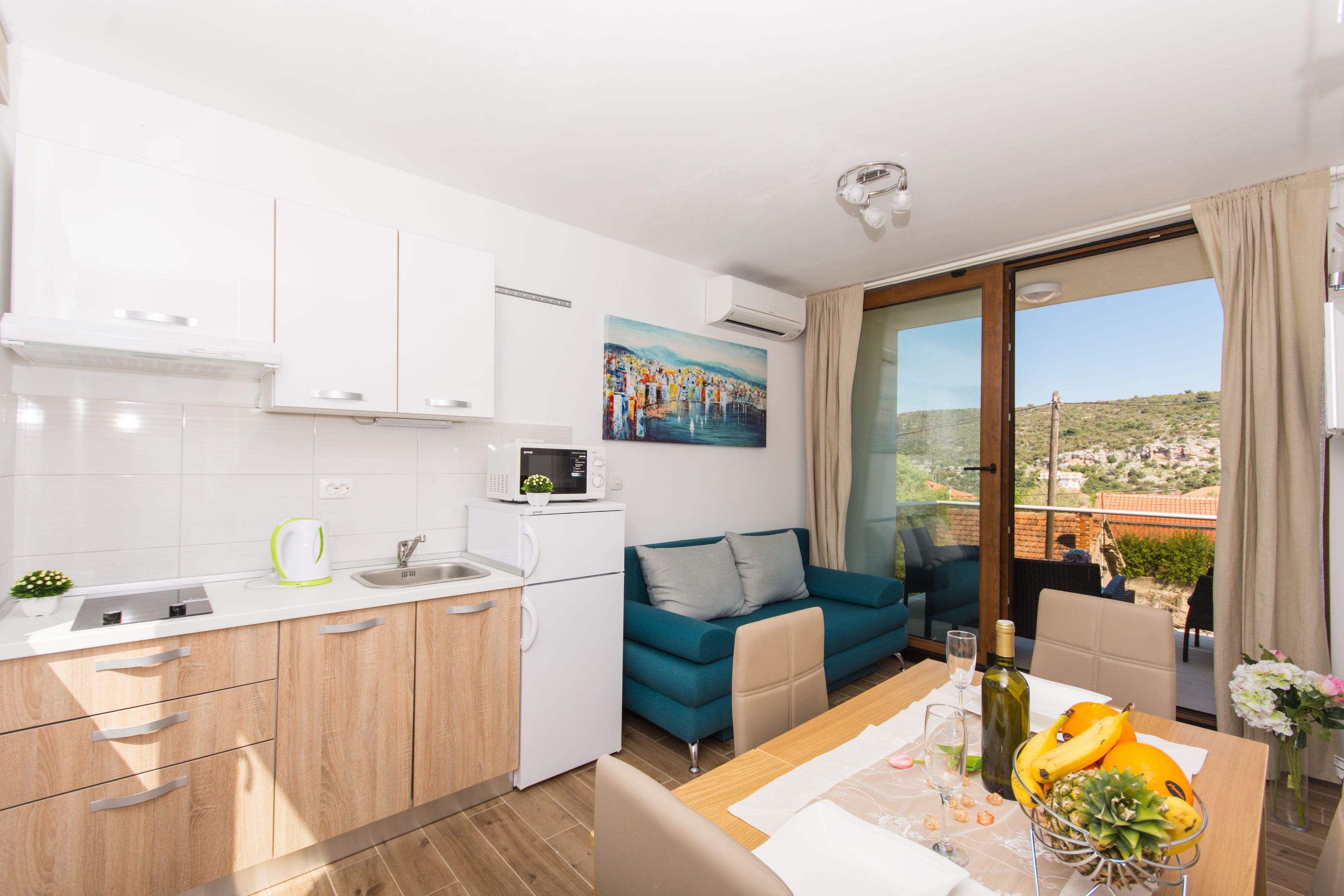 A modern kitchen with white cabinets, a microwave, and a kettle. There's a dining area with a table set for two, a blue sofa, and a view of the city through large windows.
