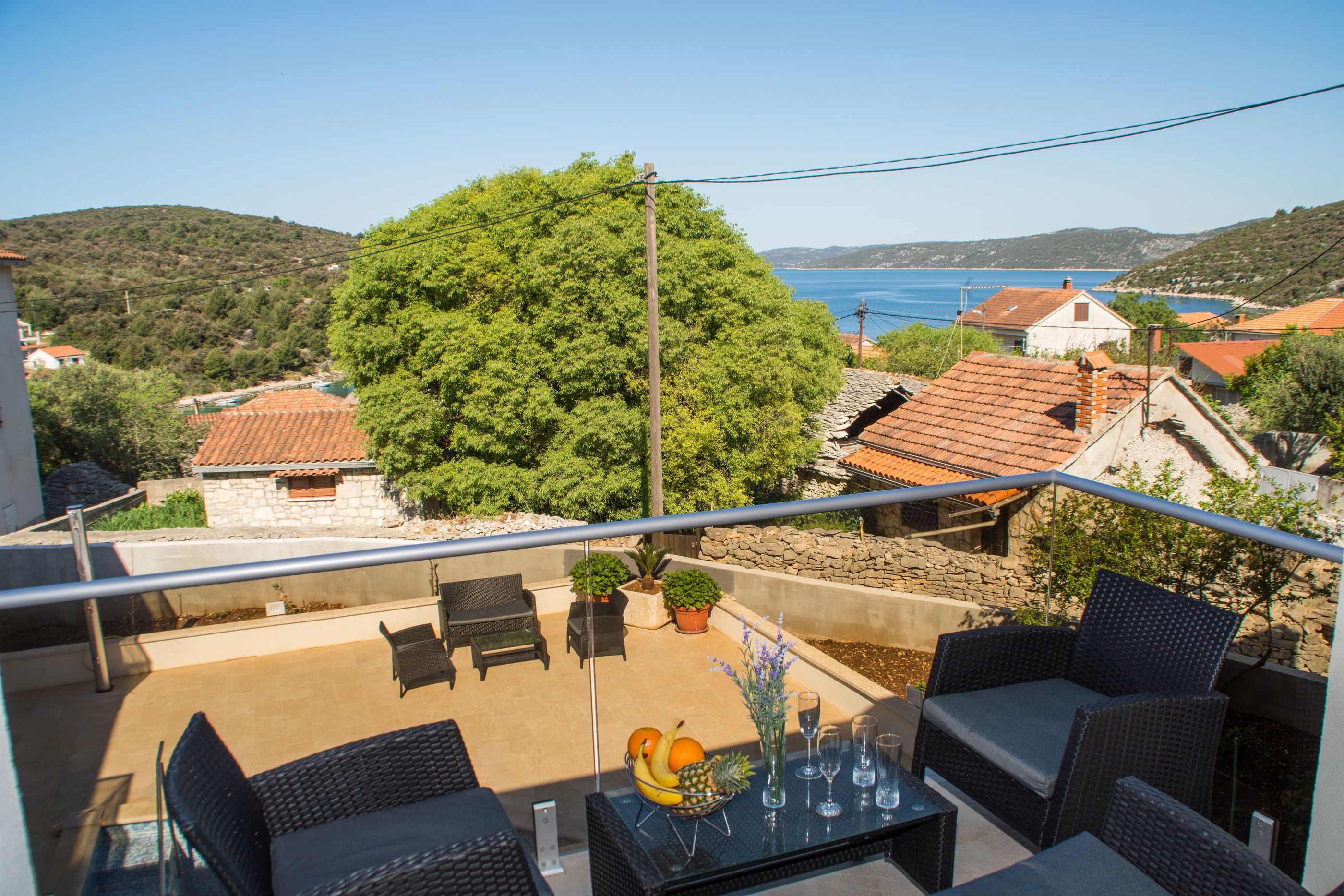 Un balcón con mobiliario de mimbre, una mesa con fruta y vasos, y vistas al mar y las colinas.