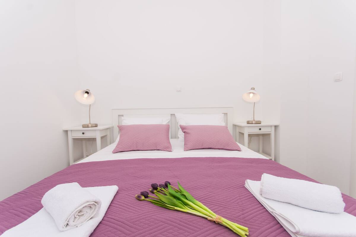 A neatly made bed with a purple quilt, white pillows, and two bedside tables with lamps.