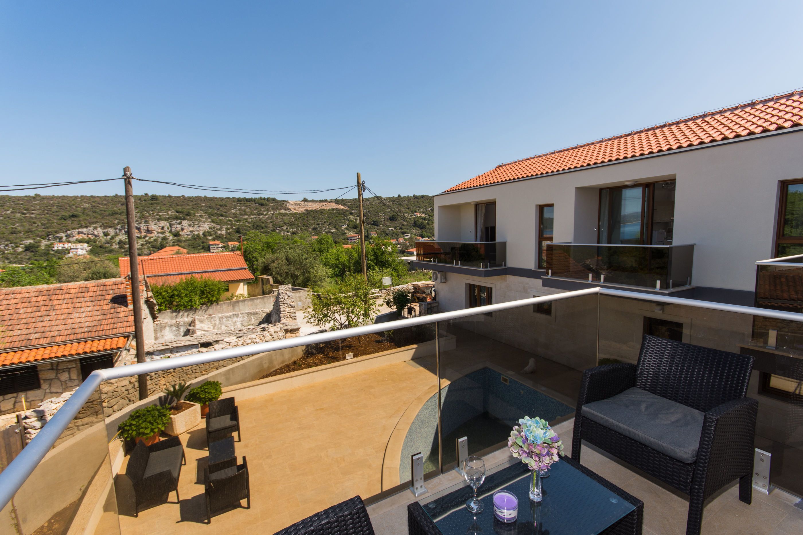 A modern outdoor balcony with a glass railing, wicker furniture, and a view of the surrounding landscape.