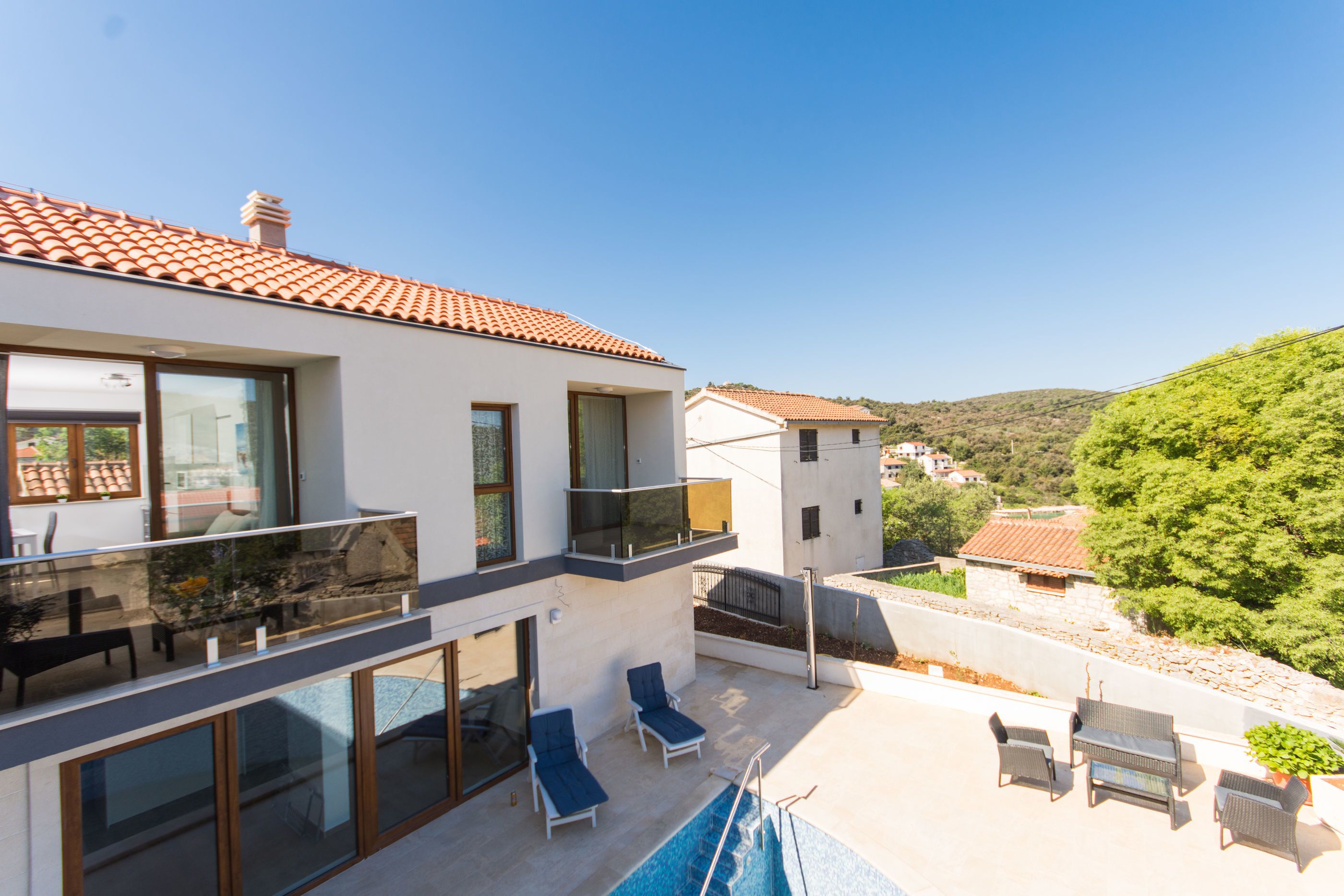 A modern two-story house with a balcony, a swimming pool, and lounge chairs.