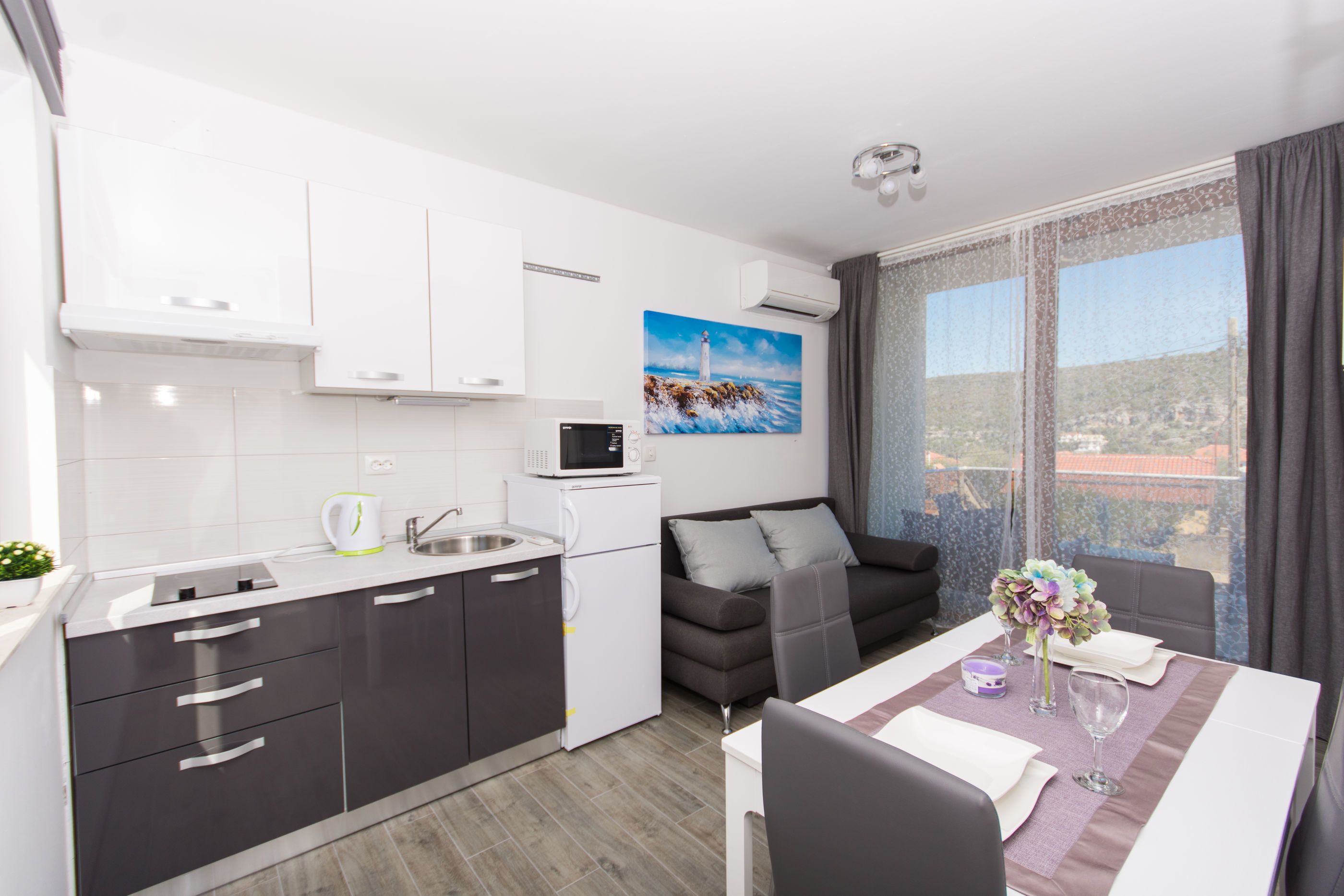 Cocina moderna con armarios blancos, microondas y cafetera. Hay un comedor con una mesa preparada para dos personas, un sofá y un ventanal con vistas al paisaje.