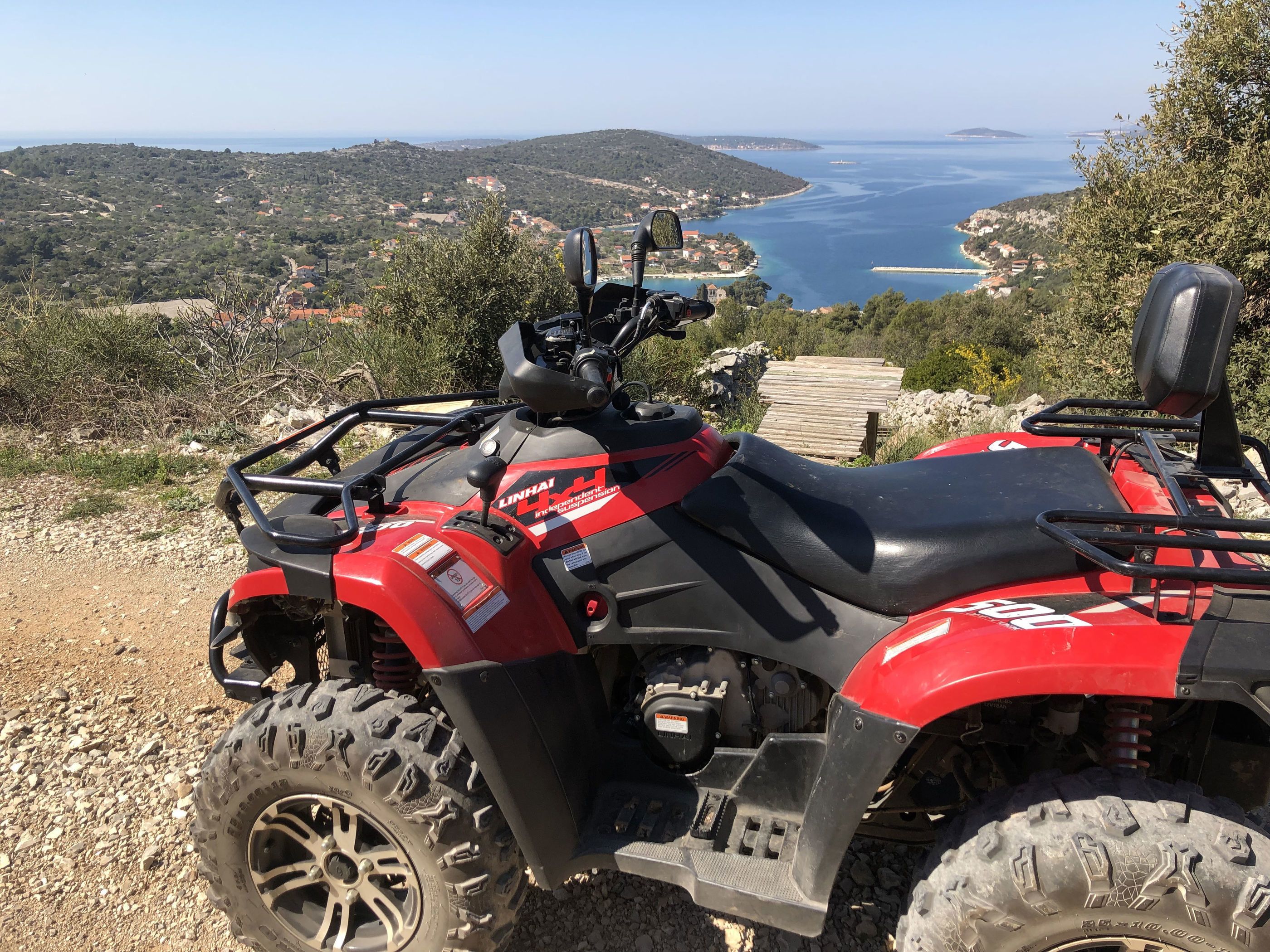 Un véhicule tout-terrain rouge et noir est garé sur un terrain rocheux, offrant une vue panoramique sur un paysage côtier en arrière-plan.