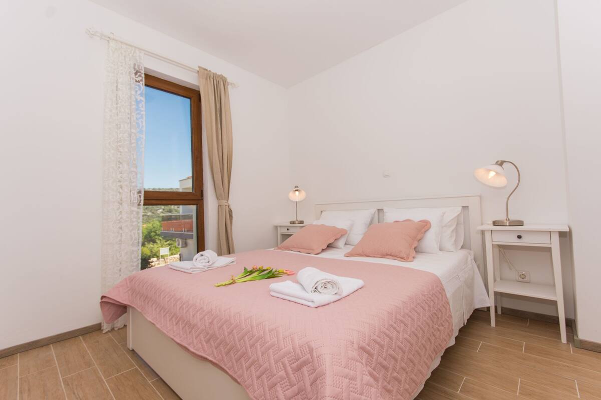 A bedroom with a bed, two bedside tables, a window with curtains, and a lamp on one table.