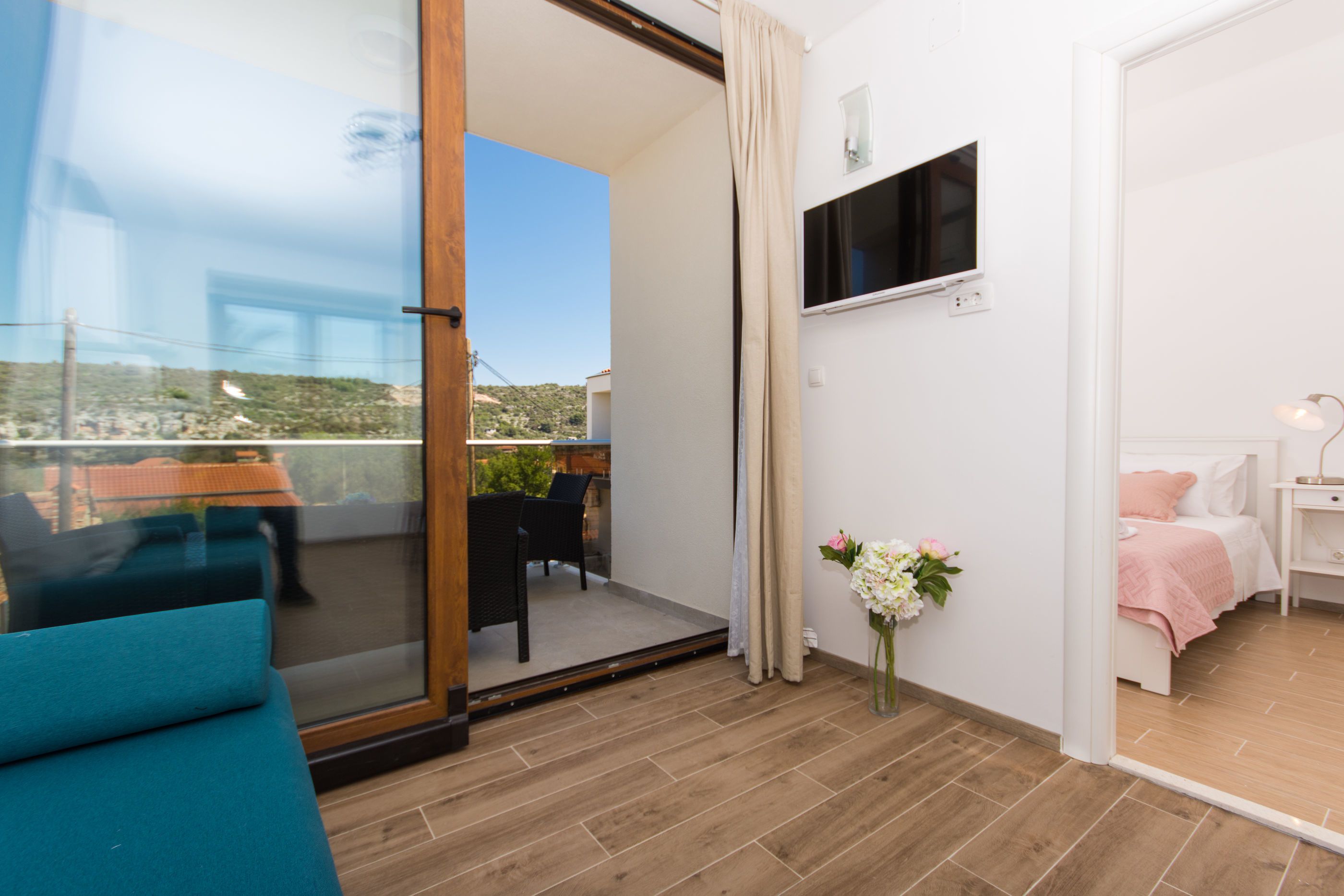 A balcony with a glass door, a TV mounted on the wall, a bed with pink bedding, and a small table with a lamp.