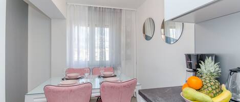 A modern dining area with a glass table, pink chairs, and a fruit bowl on the counter.