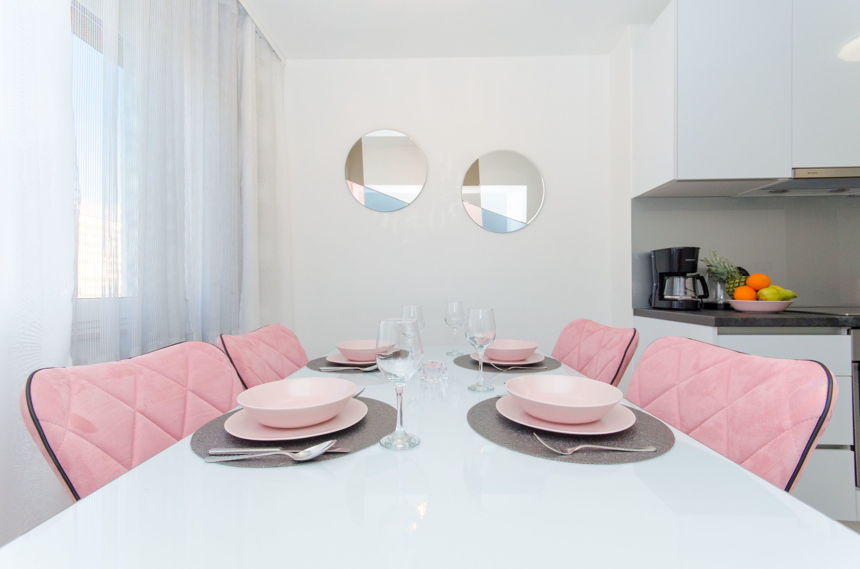 Un comedor con una mesa blanca dispuesta para seis personas, sillas rosadas y una encimera de cocina con cafetera y frutero.