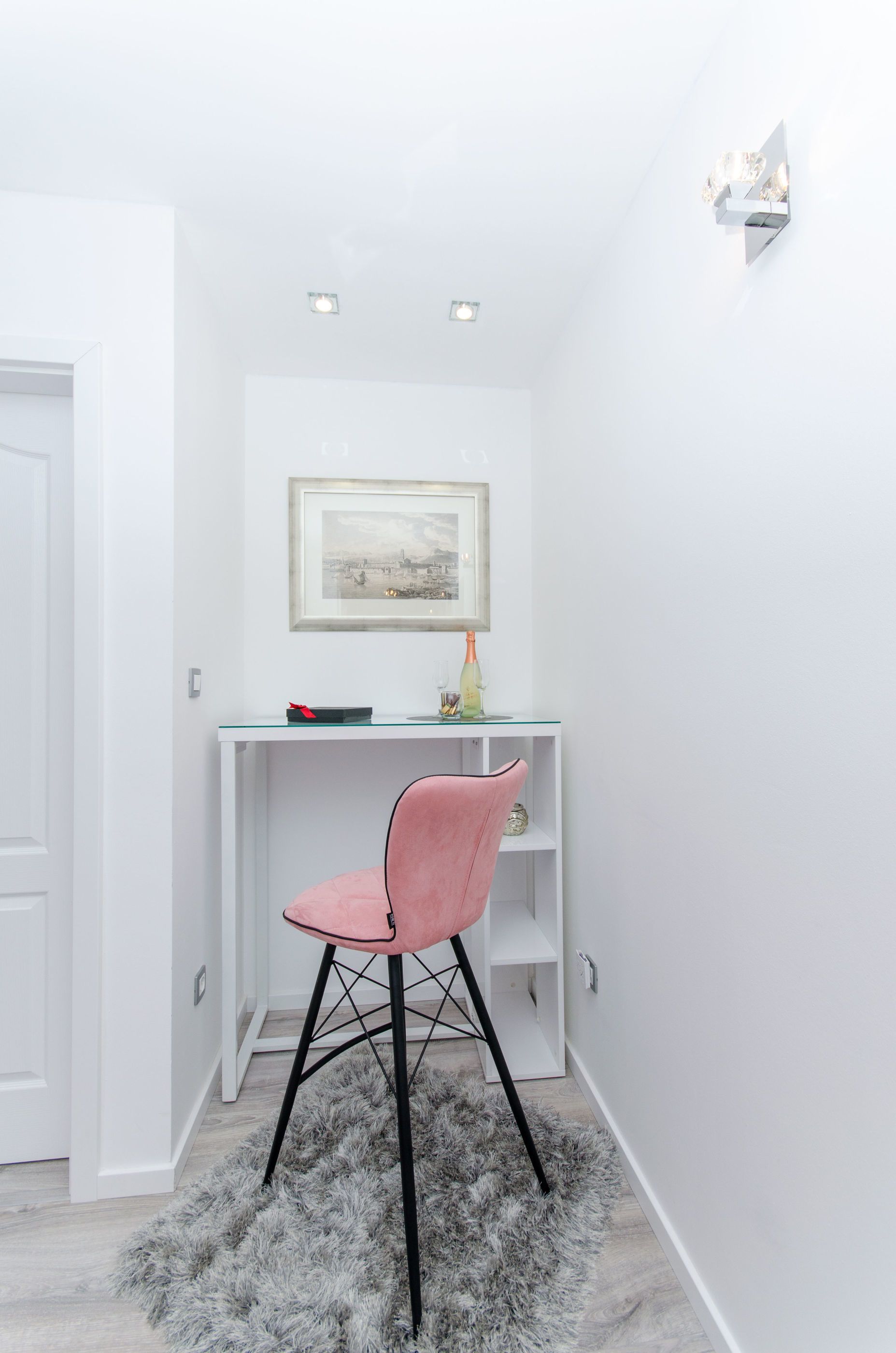 A small, modern room with a white desk, a pink chair, a framed picture on the wall, and a fluffy rug on the floor.