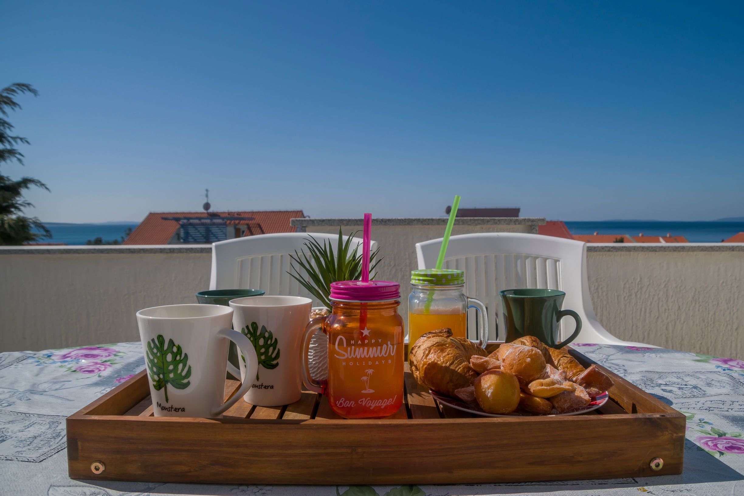 Una bandeja de madera con un desayuno que incluye tazas de café, un tarro de jugo de naranja, una taza verde y pasteles sobre una mesa con vista al mar y edificios.