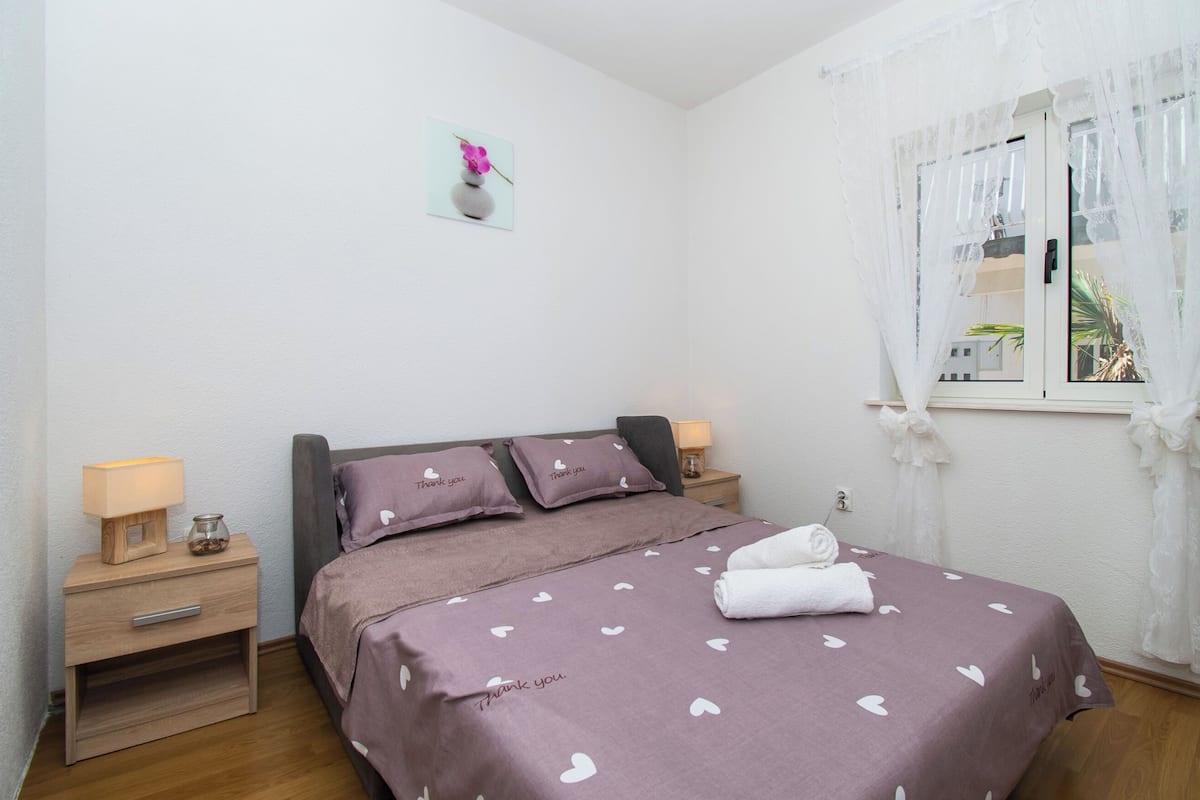 Ein ordentlich eingerichtetes Schlafzimmer mit einem Bett, zwei Nachttischen, einem Fenster mit durchsichtigen Vorhängen und einem Wandbild.