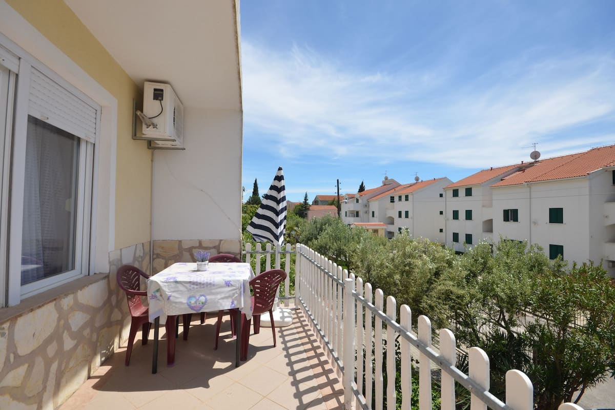 Ein Balkon mit Tisch und Stühlen, einem gestreiften Sonnenschirm und Blick auf Gebäude im Hintergrund.