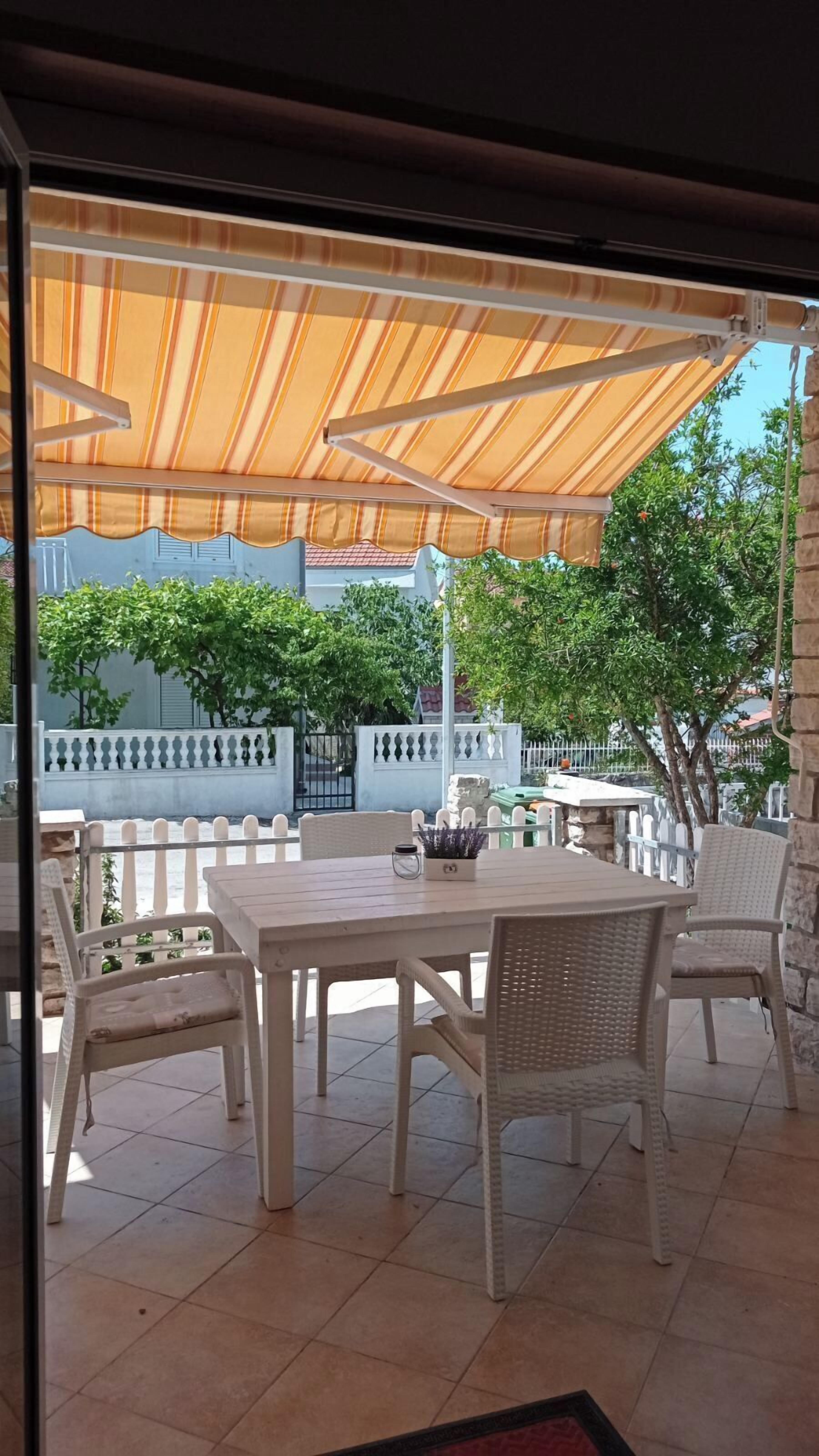Une terrasse extérieure avec une table et des chaises, un auvent à rayures et une vue sur un jardin et des bâtiments en arrière-plan.