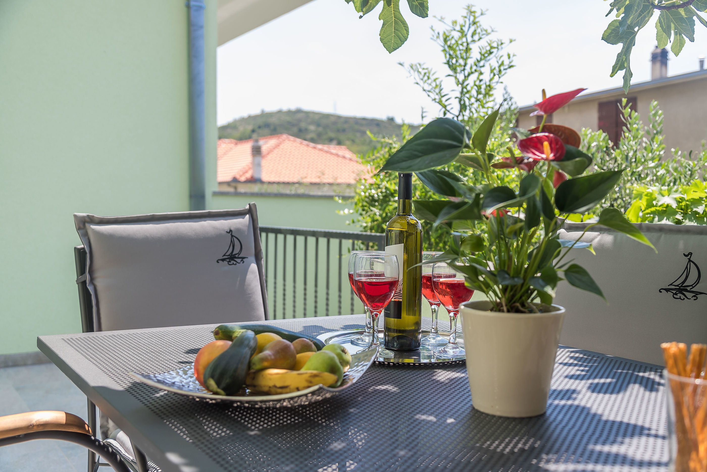 Una mesa con fruteros, copas de vino y una botella en un balcón con vista a un edificio y follaje.