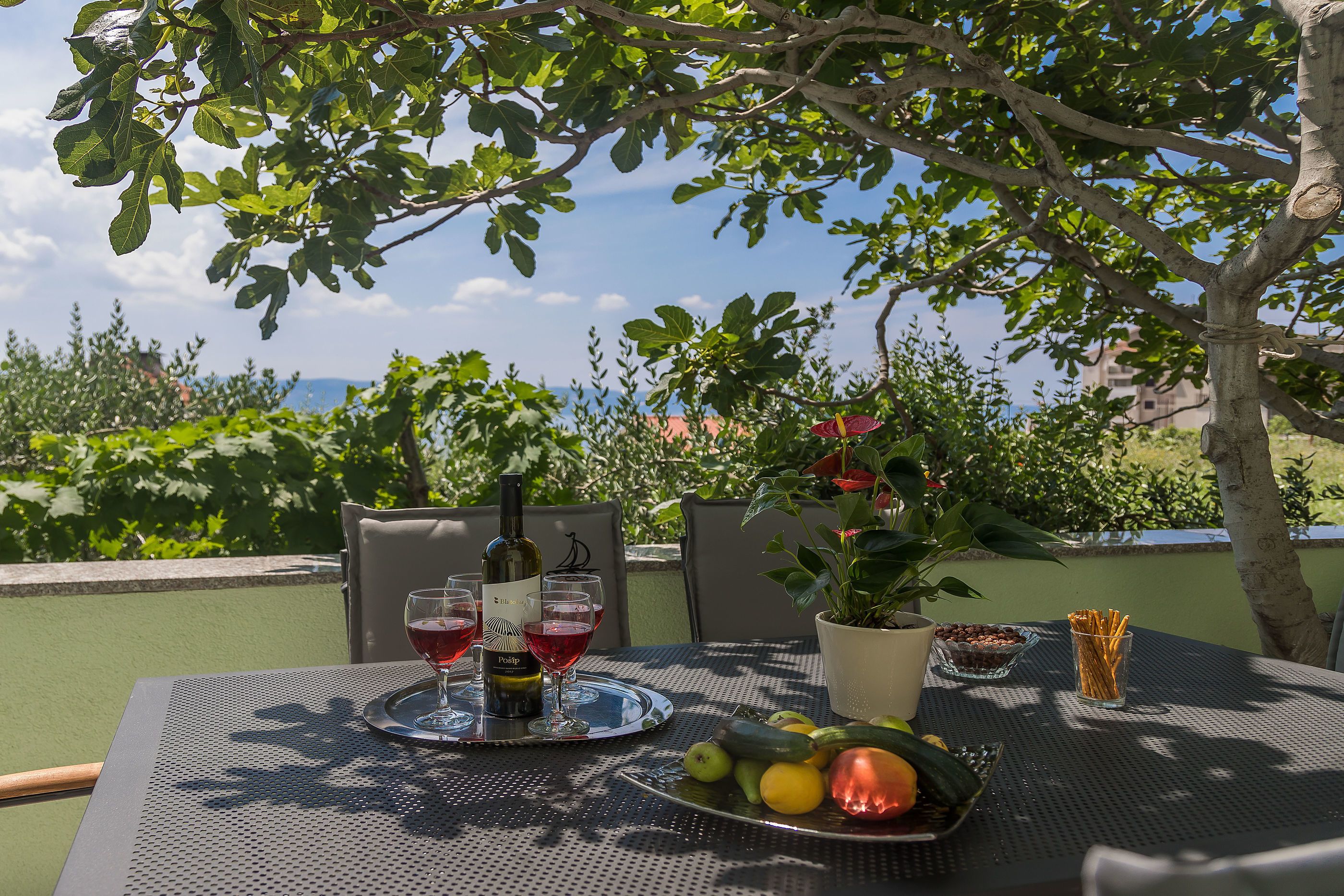 Une table à manger extérieure avec une bouteille de vin, des verres et une assiette de fruits.