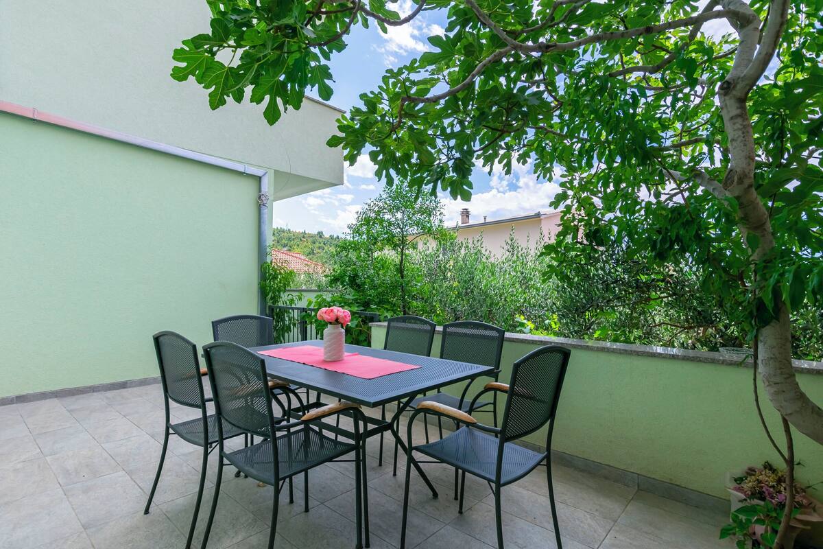 An outdoor patio with a table and chairs, surrounded by greenery and a clear sky.